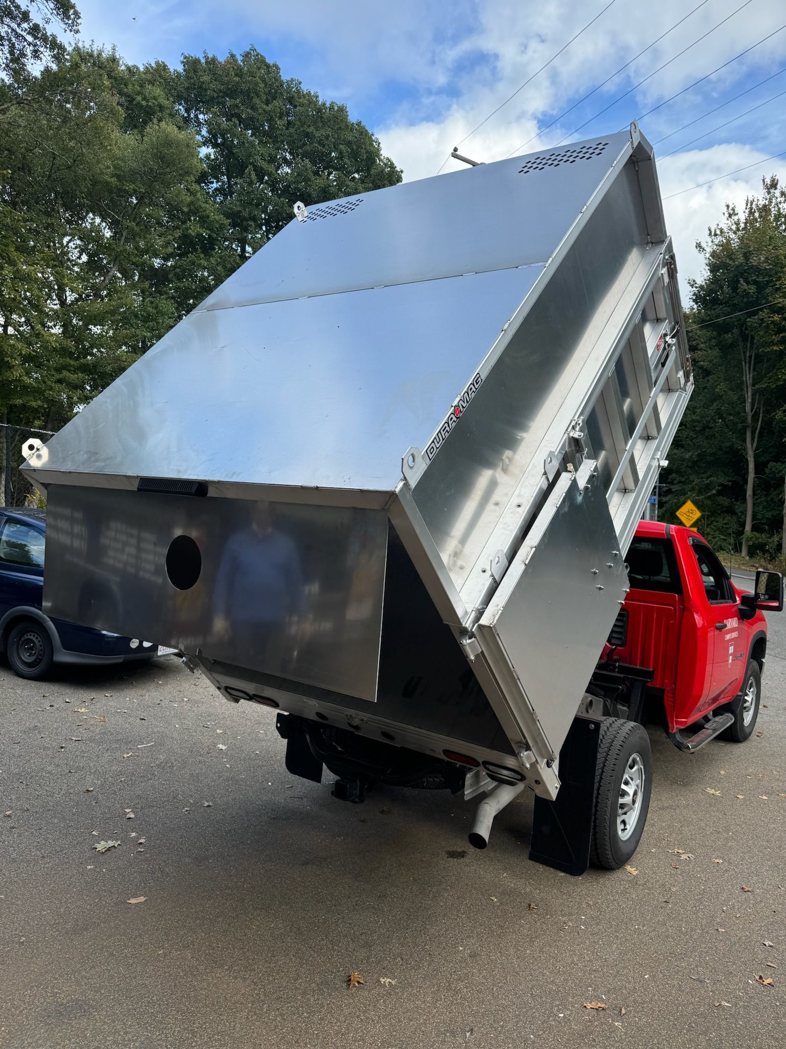 Shiny metal dump trailer tilted steeply on a red utility vehicle in a wooded parking area.