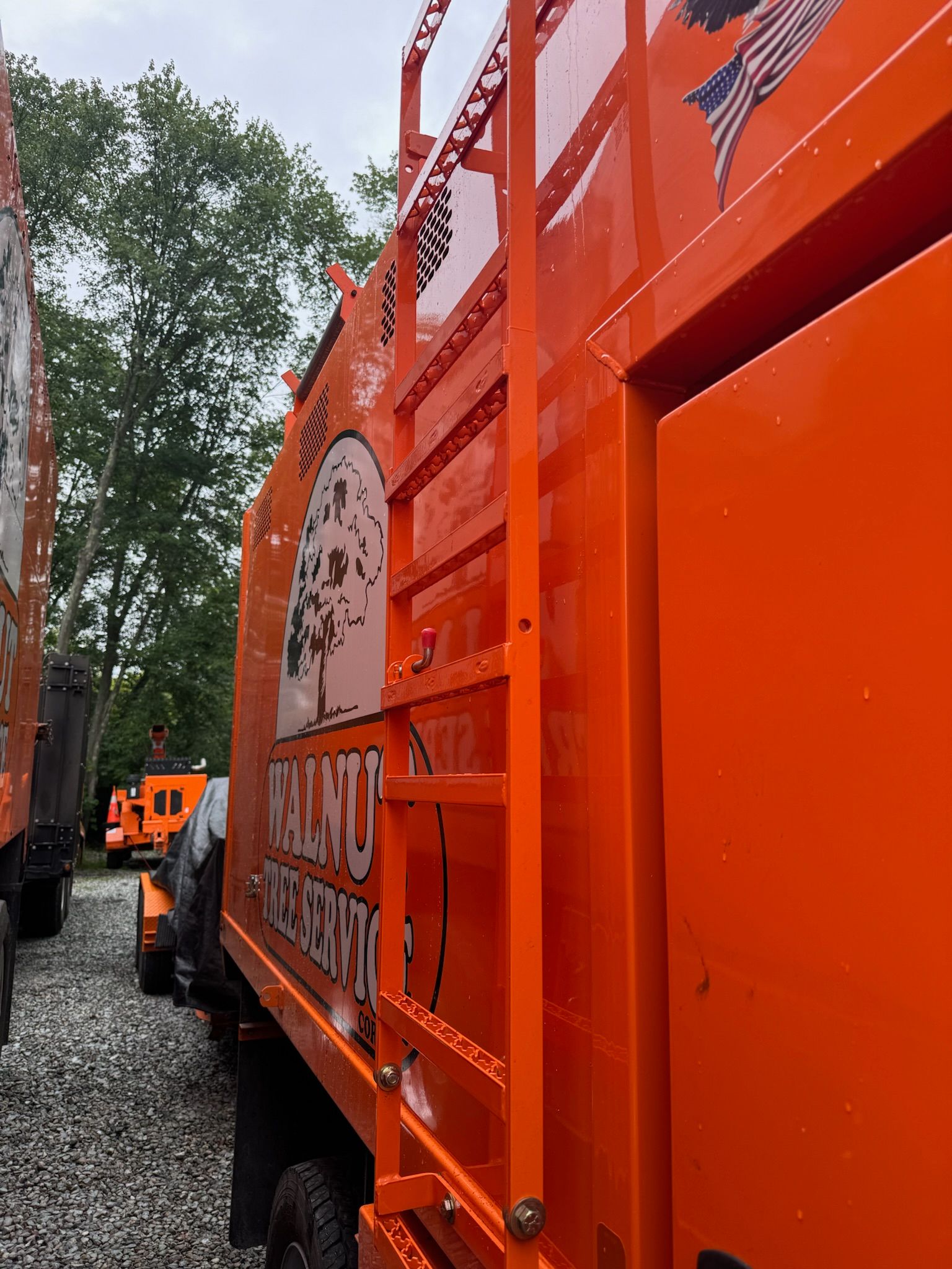 Orange garbage truck in a gravel lot, viewed from the side with trees in the background.