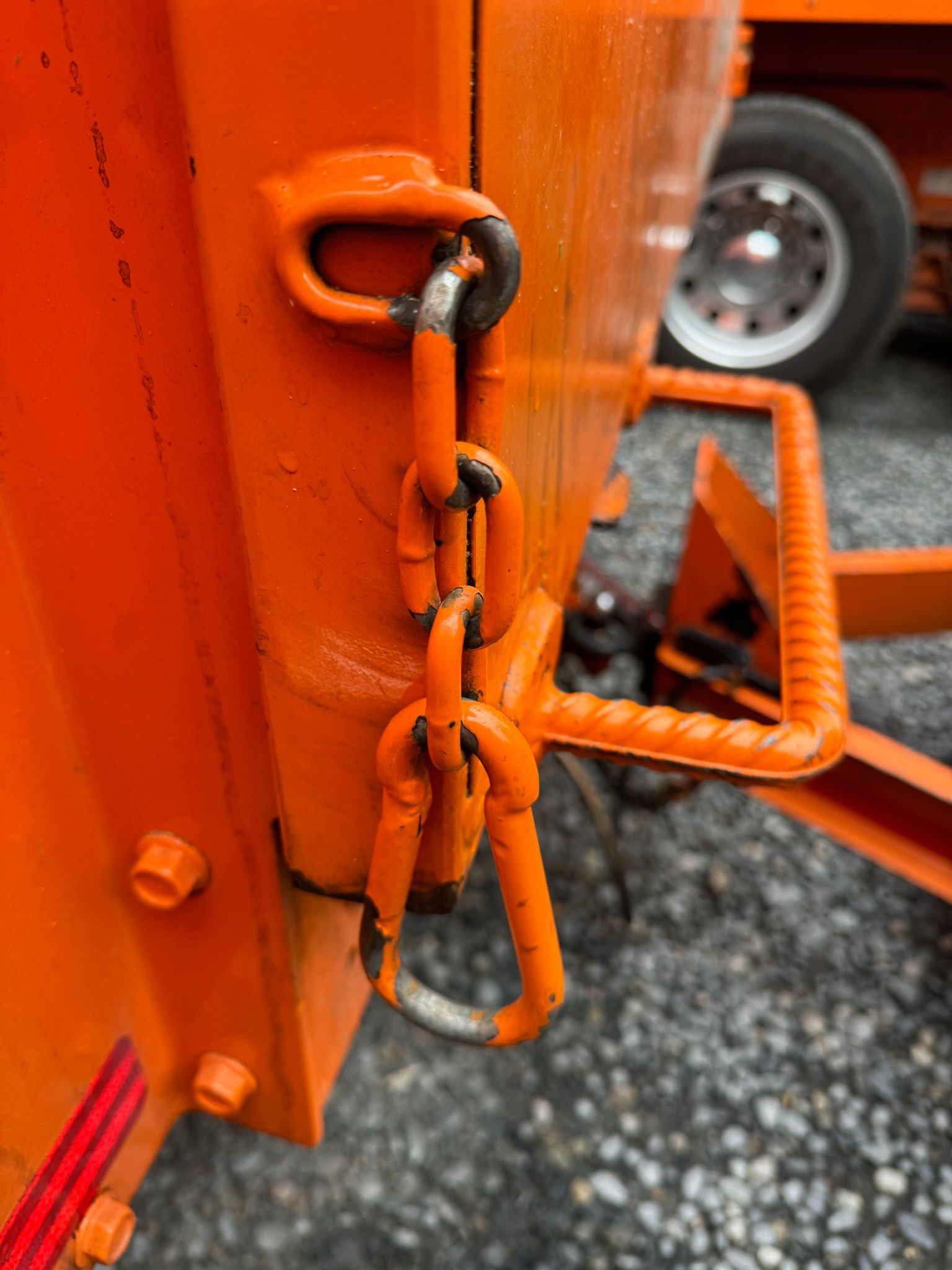 Orange trailer latch secured with a chain, shown close-up beside a wheel on gravel.