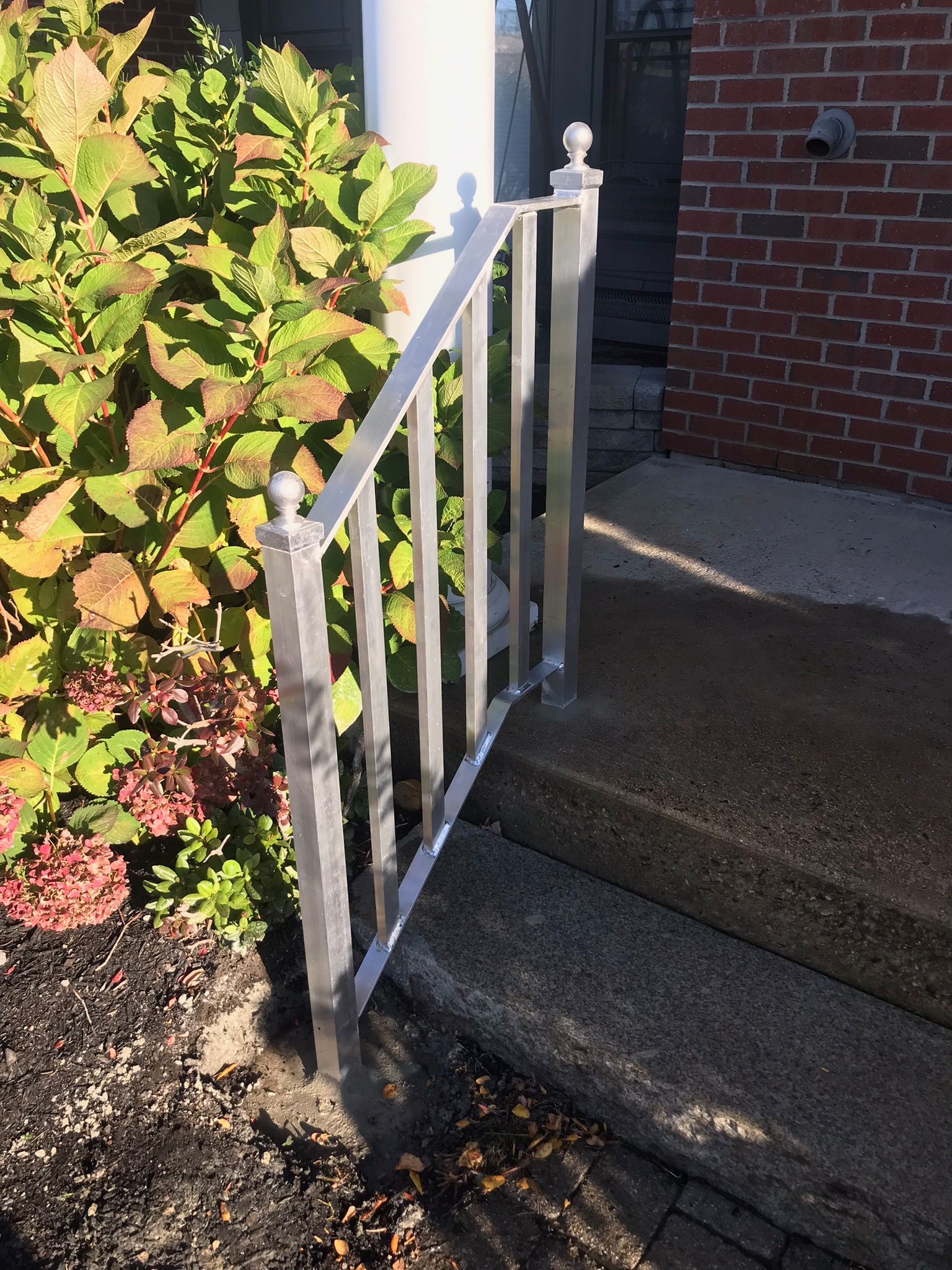 White metal handrail beside front steps and walkway next to a brick house and green shrubbery