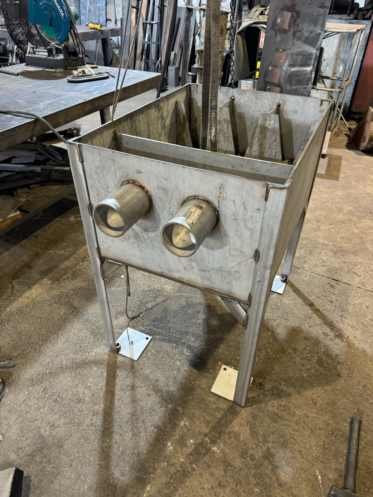 Metal hopper on four legs with two pipe fittings, standing on a workshop floor.