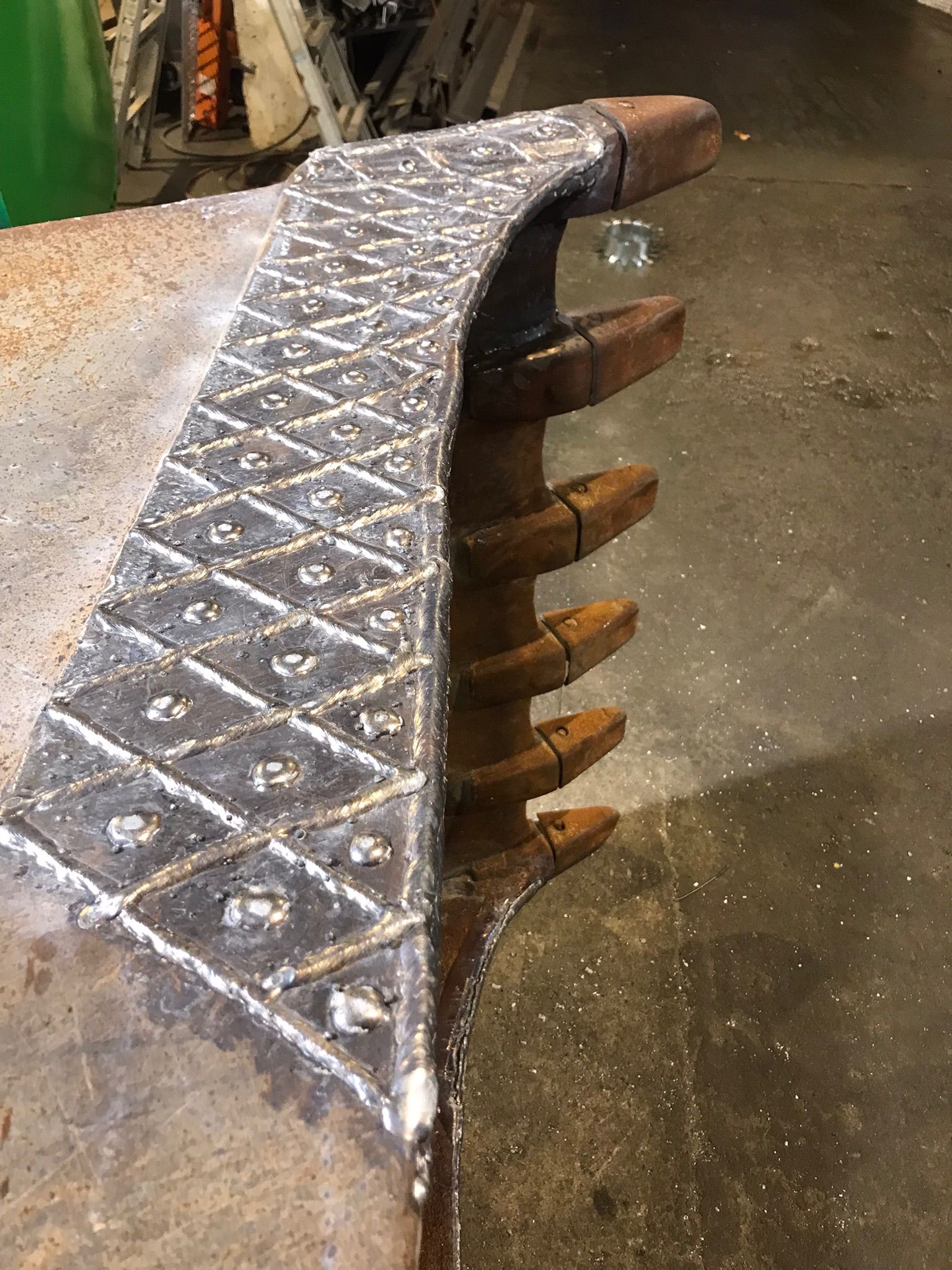 Close-up of a metal excavator bucket with worn, welded teeth on a concrete floor