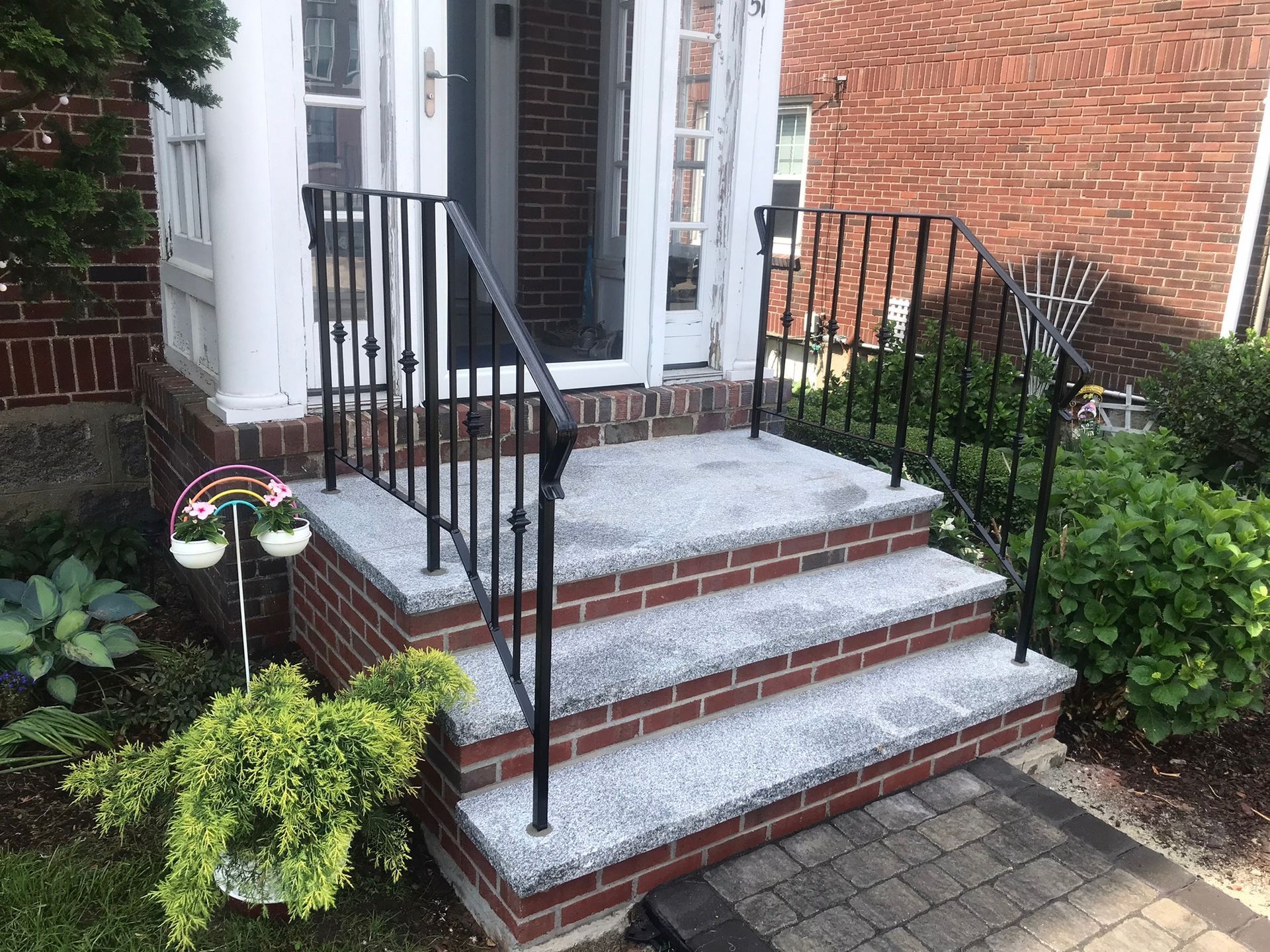 Brick front steps with black railings leading to a white door, bordered by flower pots and shrubs.