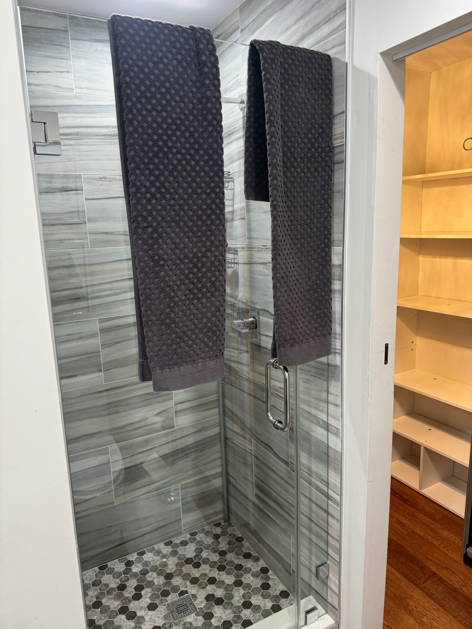Narrow tiled shower with mosaic floor, gray wall tiles, and dark hanging towels or shower curtains on both sides