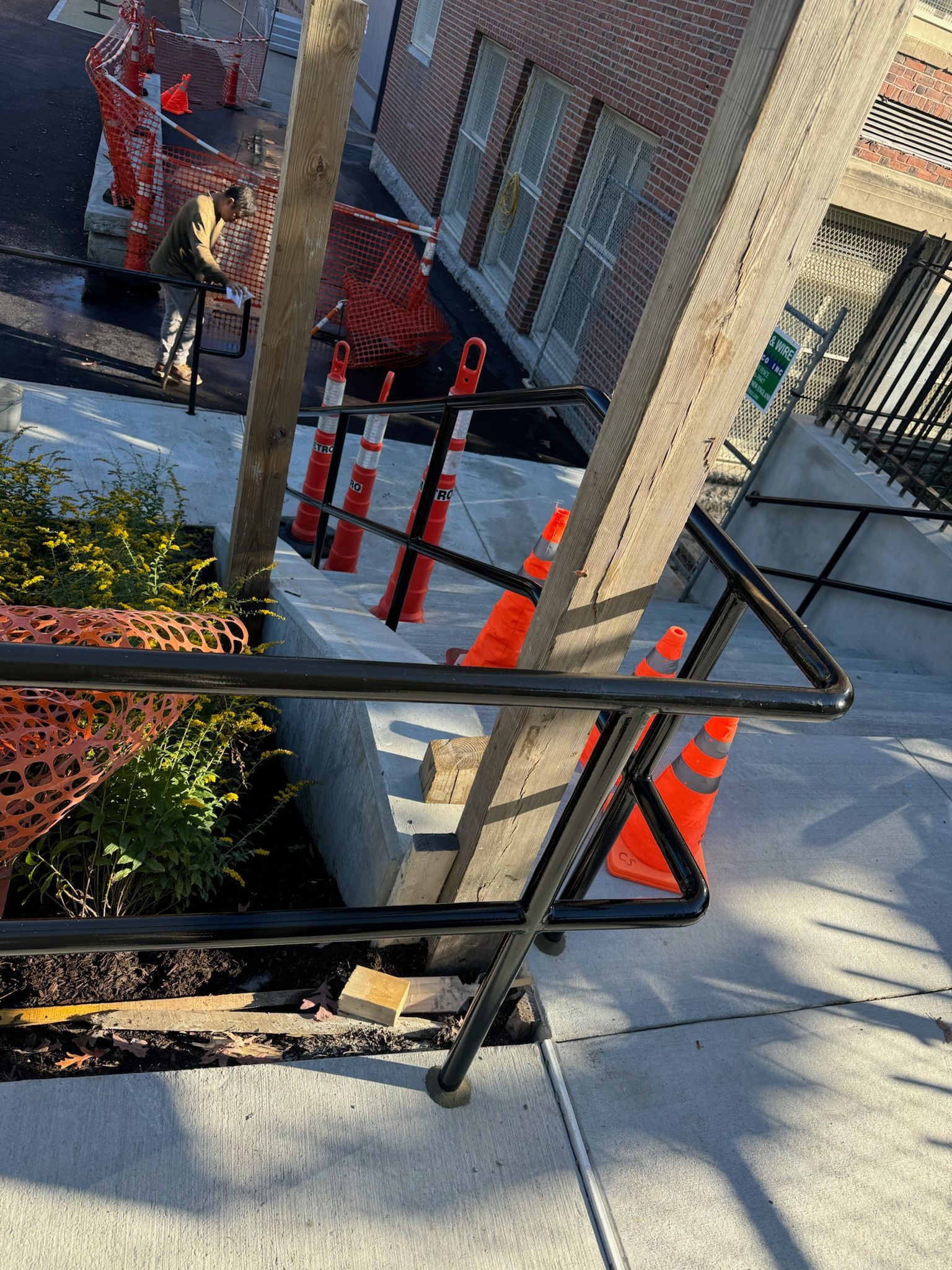City sidewalk with black railings, orange cones, and a brick building entrance by a planted bed.