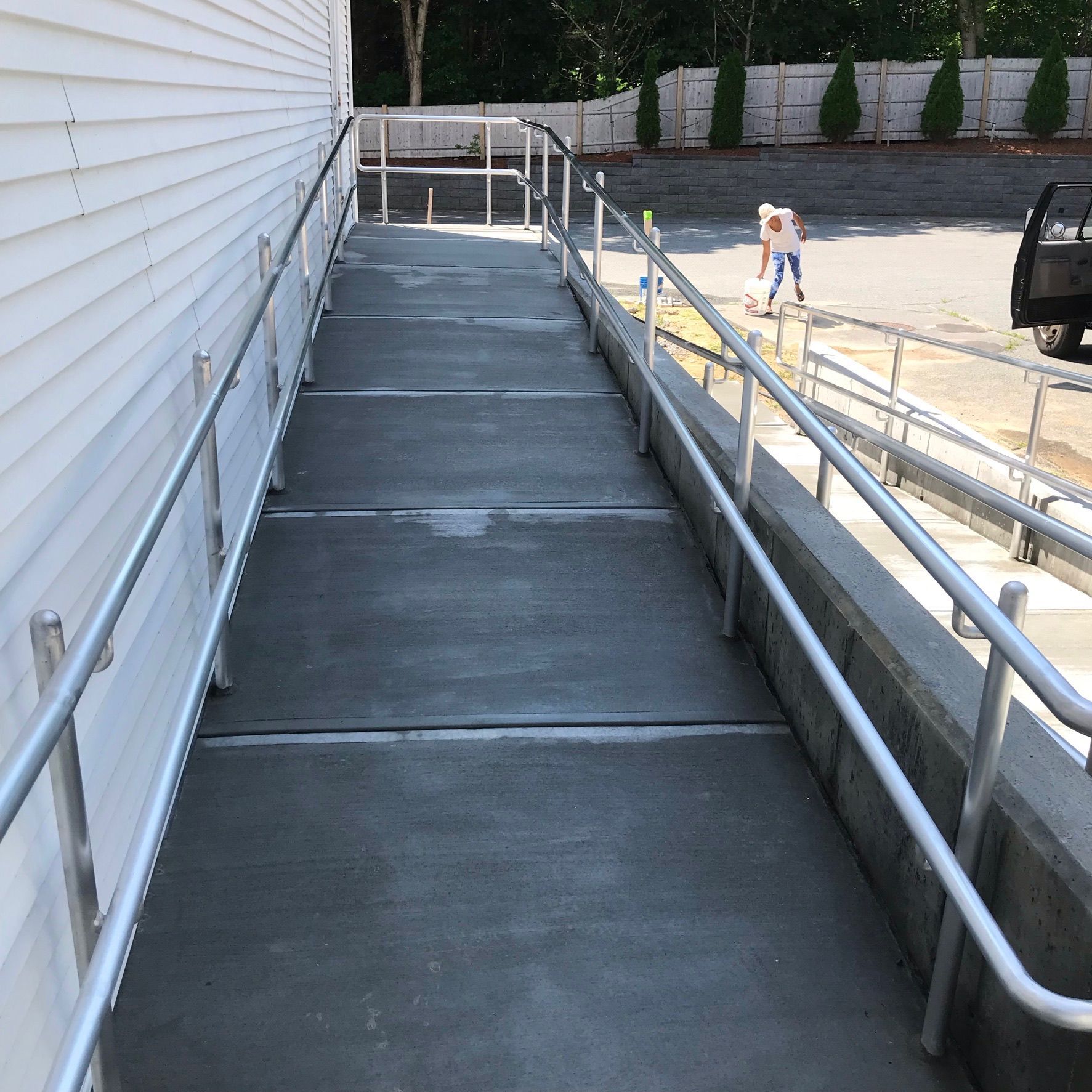 Concrete accessibility ramp beside a building, with white railings and a parking lot in the background