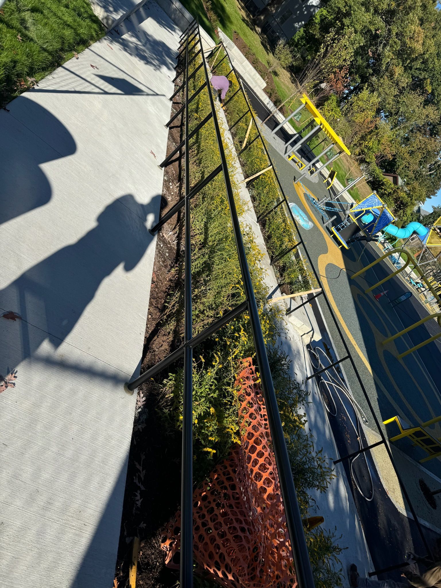 Steep outdoor walkway beside a metal fence, with trees, shadows, and orange netting below.