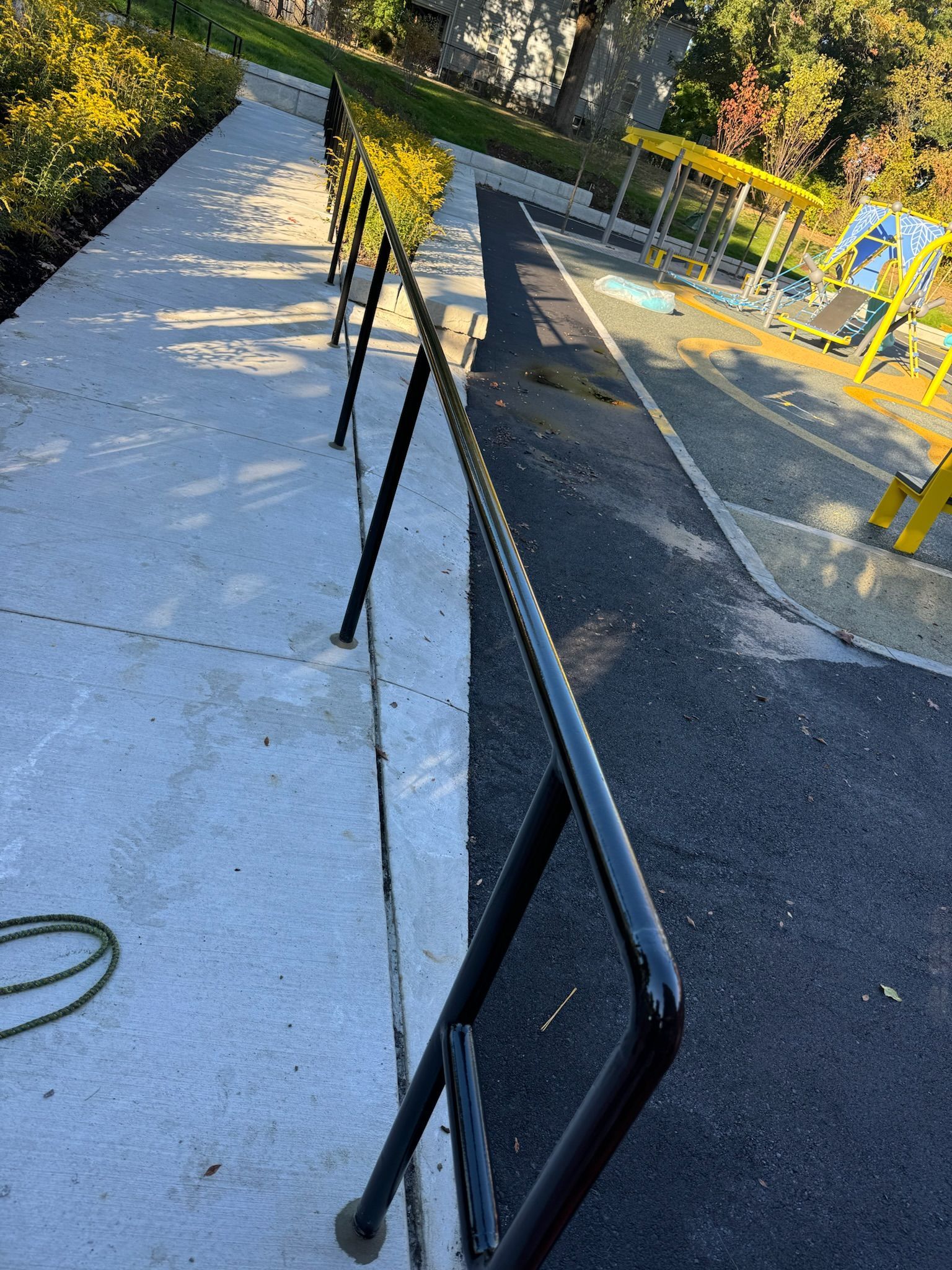 Outdoor walkway with black handrail beside a concrete path and yellow playground equipment.