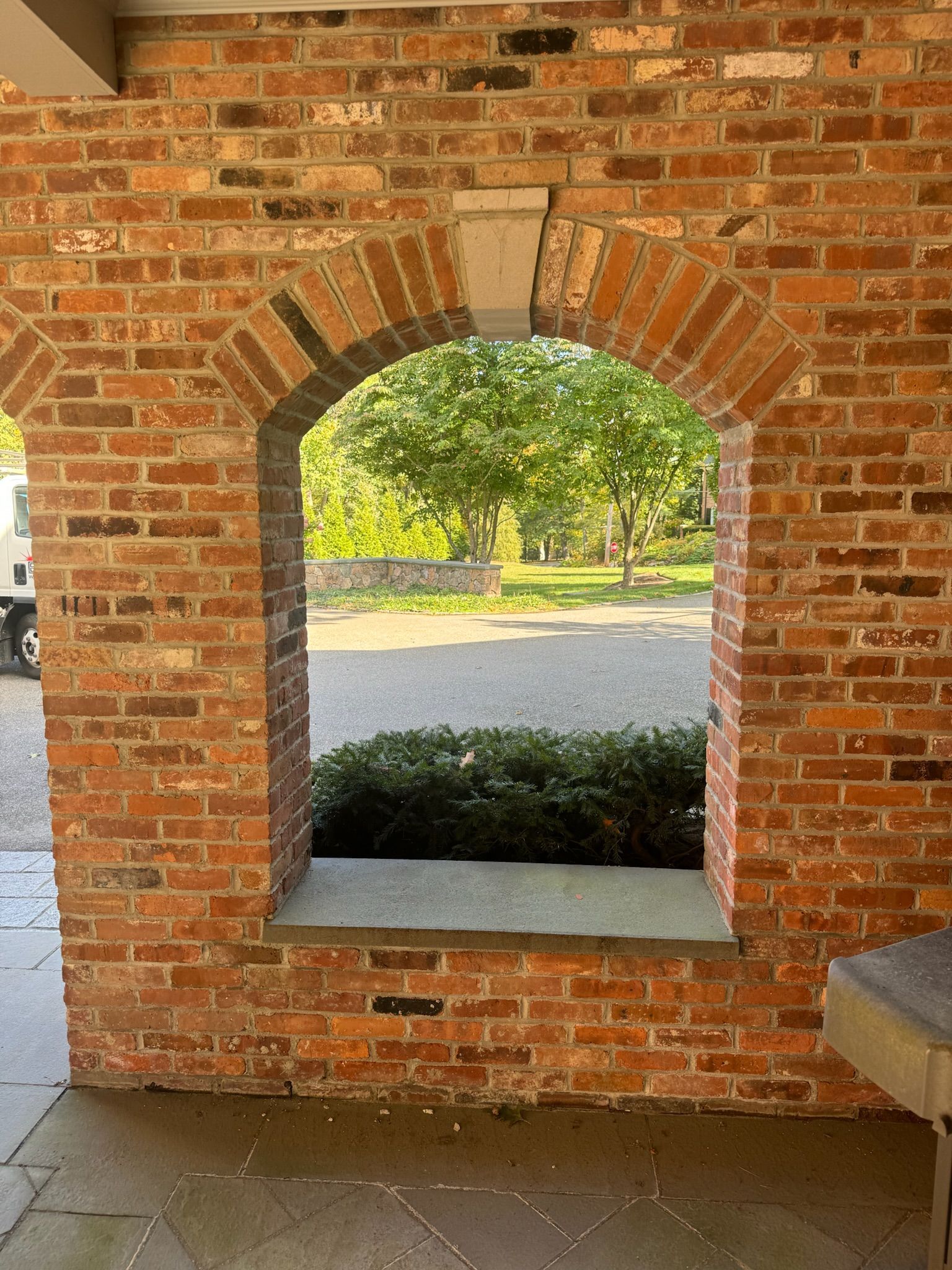 Brick arch window opening onto a sunlit park with trees and a gravel path
