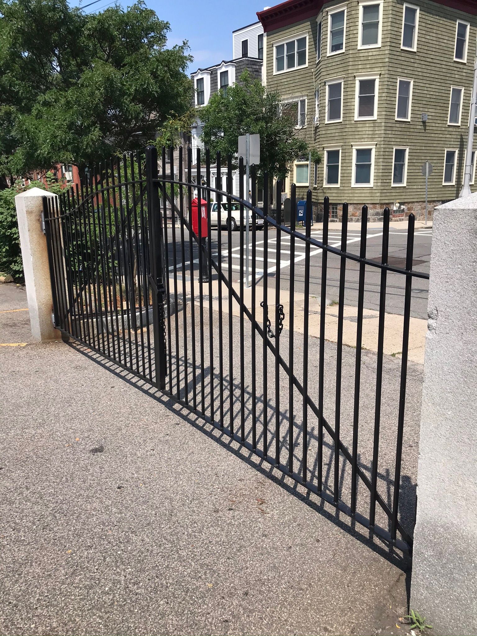Black metal driveway gate beside a stone building on a gravel entrance, with a person in red behind it.