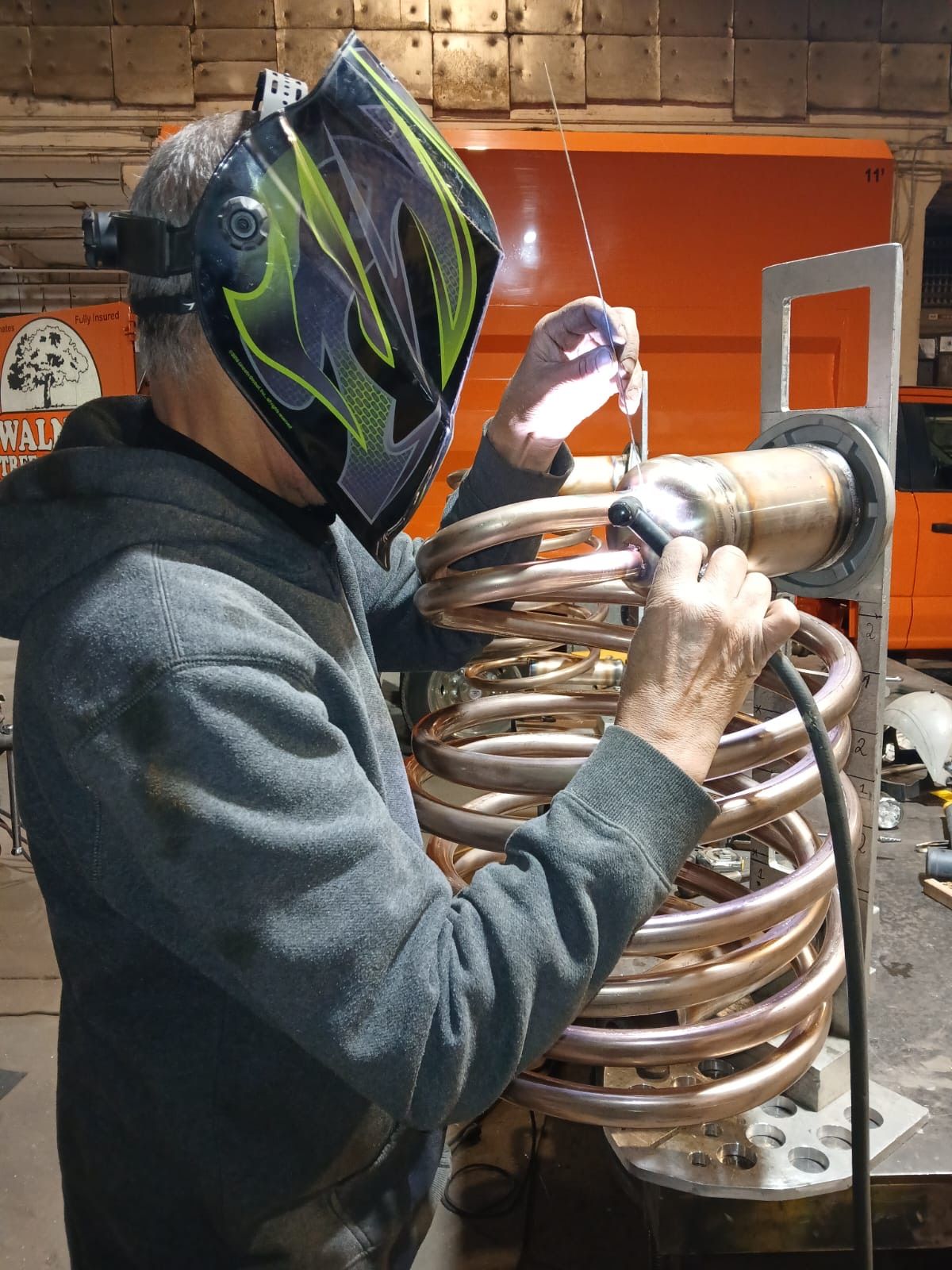 Person in welding helmet works on copper tubing inside an orange industrial machine.