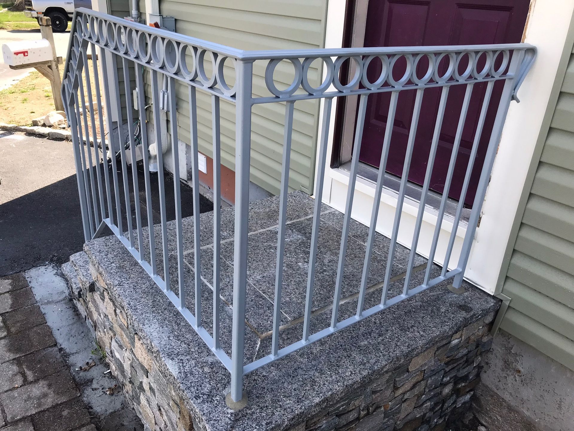 White metal railing on a small raised stone porch beside a house with beige siding and a maroon door.