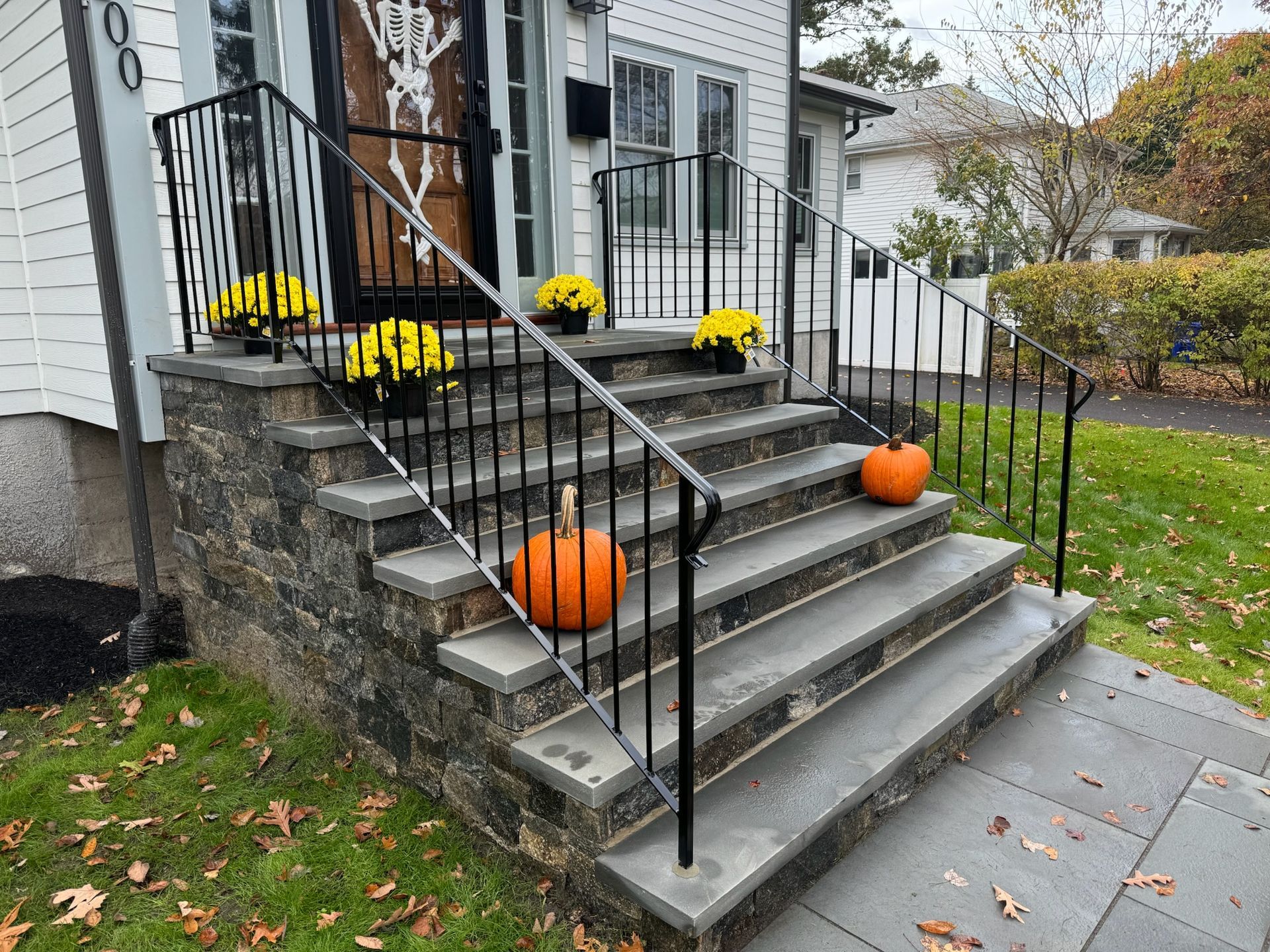 Front steps of a house with black railings, stone walls, and Halloween pumpkins and yellow flowers on the stairs
