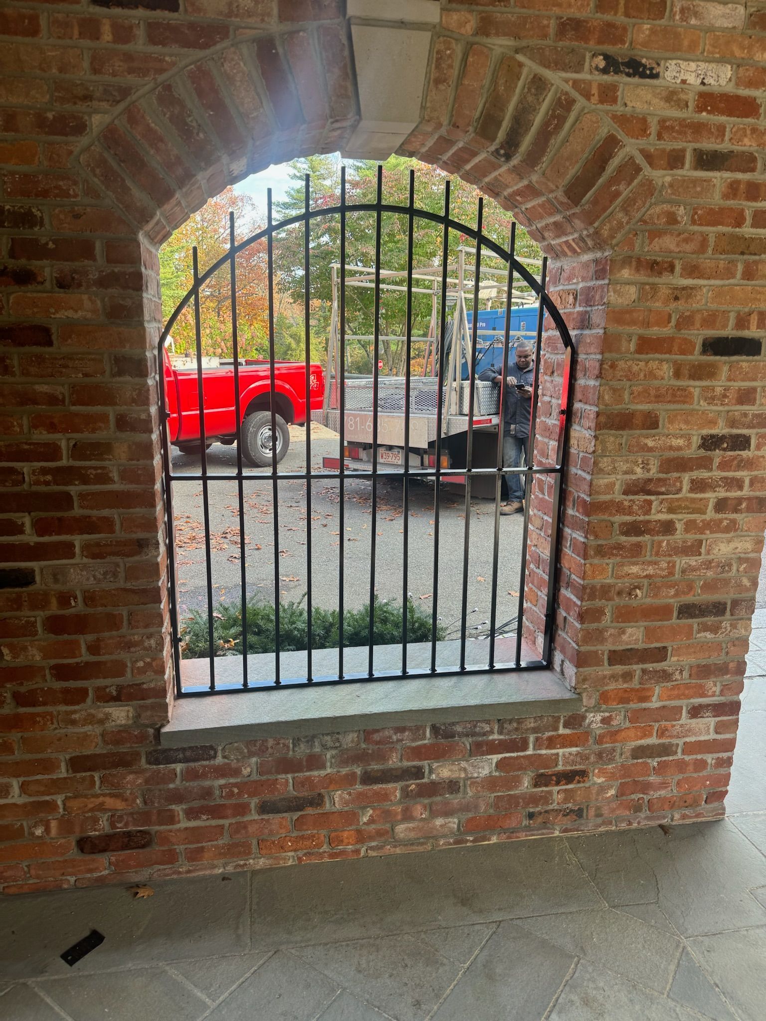 Brick arch window with iron bars overlooking a red truck and a worker outside.