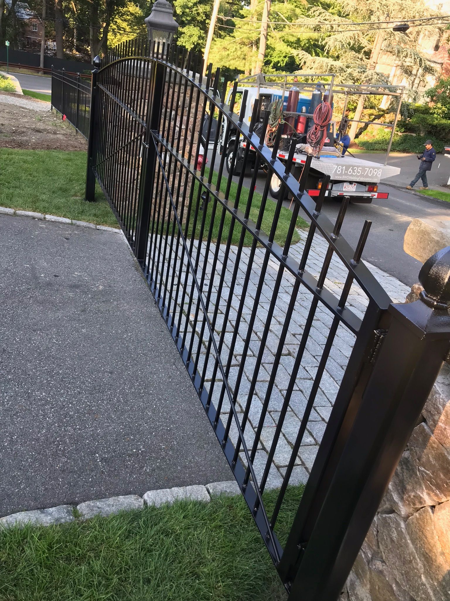 Black metal fence beside a sidewalk, with parked cars and trees along a street in the background