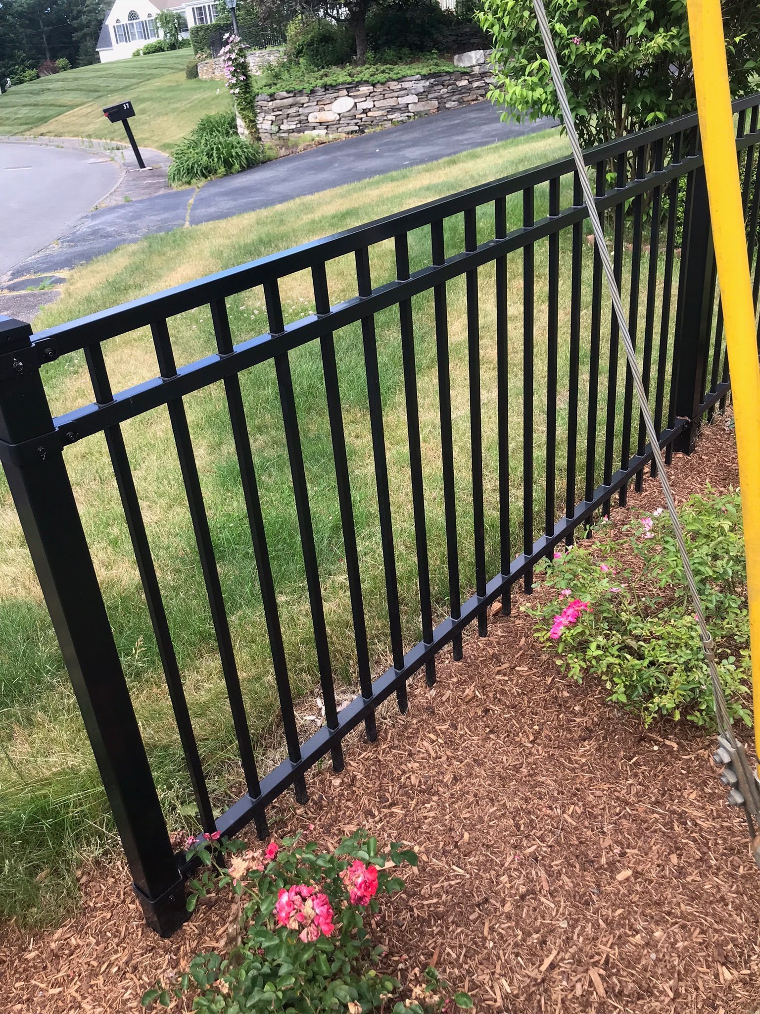 Black metal fence beside a driveway, bordered by mulch and pink flowers in a grassy yard.