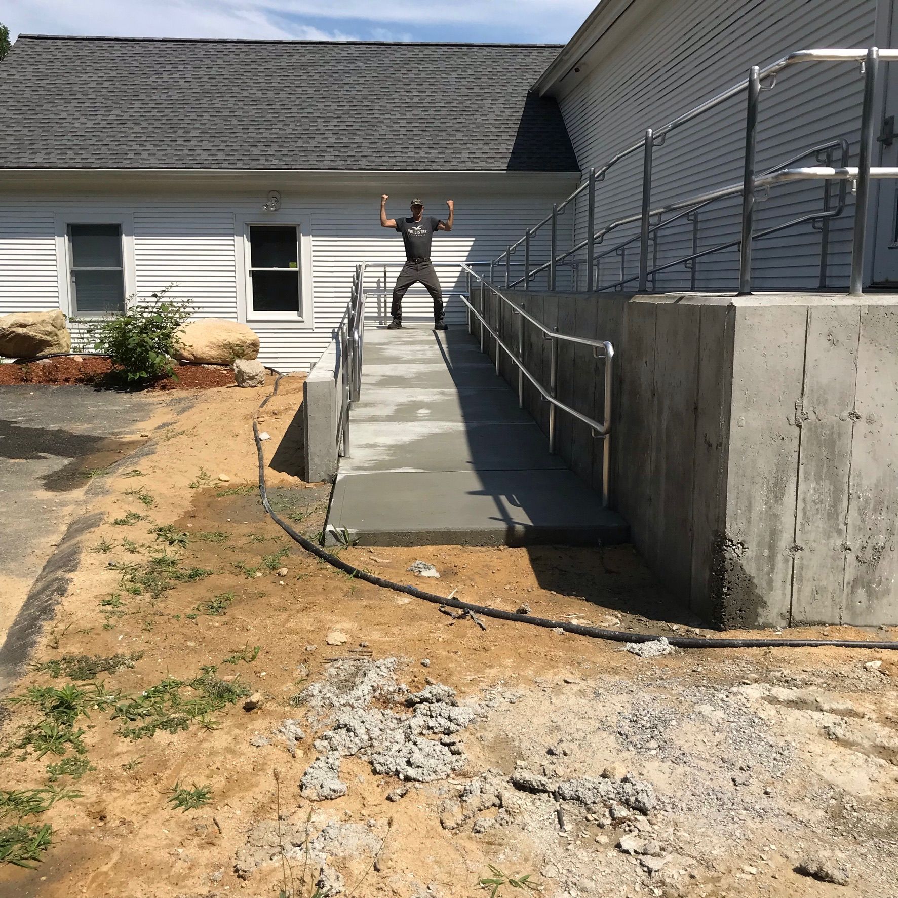 Person standing at top of a ramp beside a building and retaining wall under a bright sky