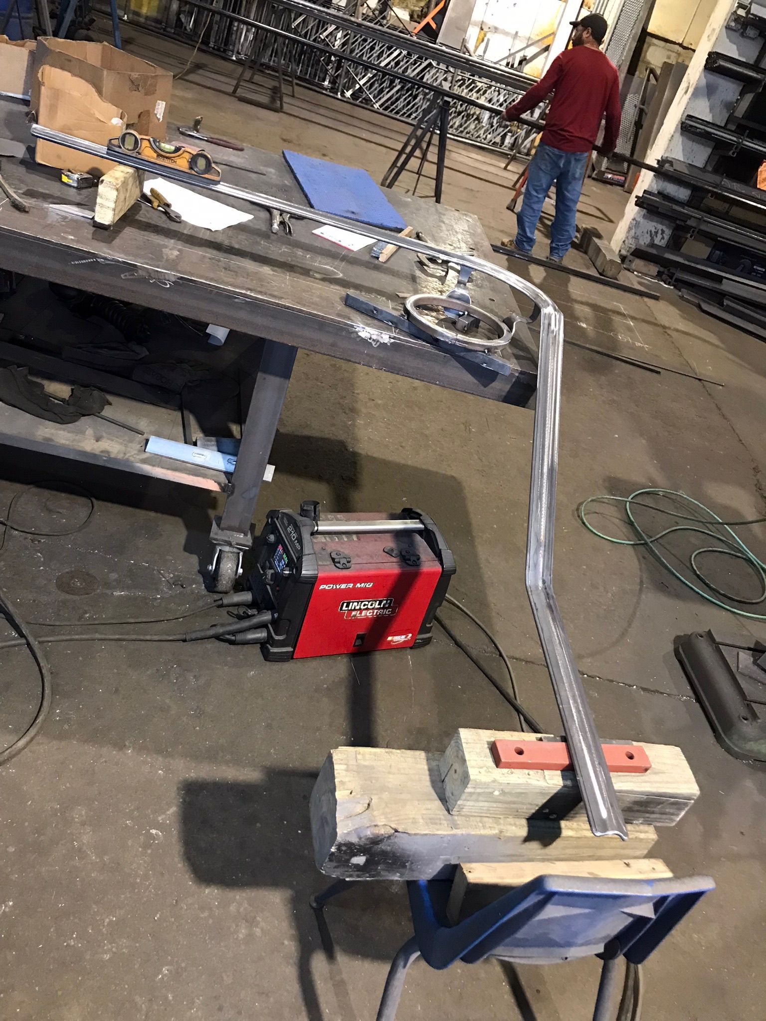 Curved metal tube clamped in a workshop vise beside a workbench and welding equipment.