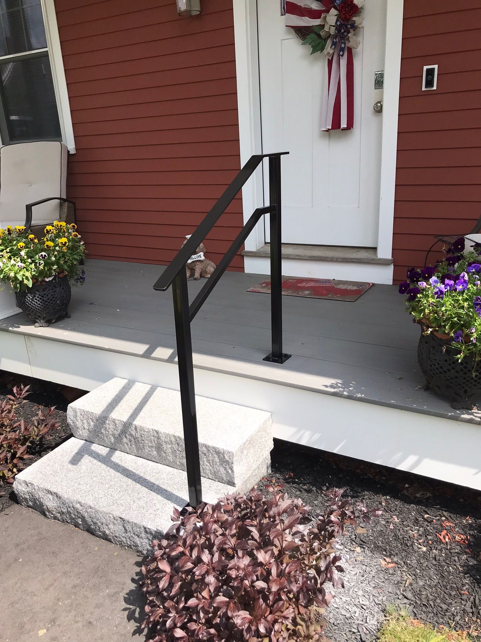 Front porch with black handrail, white door, red siding, potted flowers, and concrete steps with a small ramp