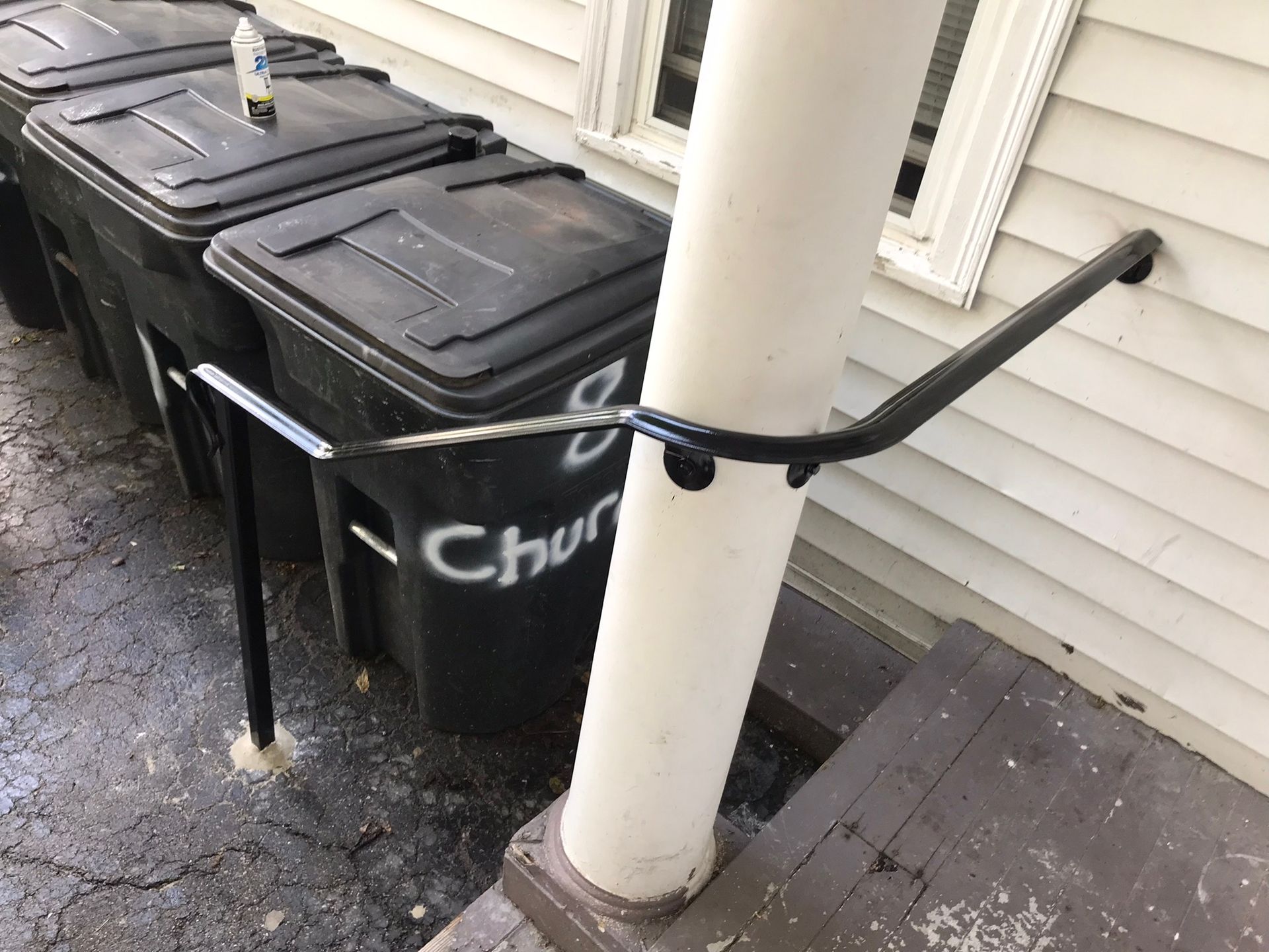 Three black trash bins beside a white porch post, with a black railing across them.