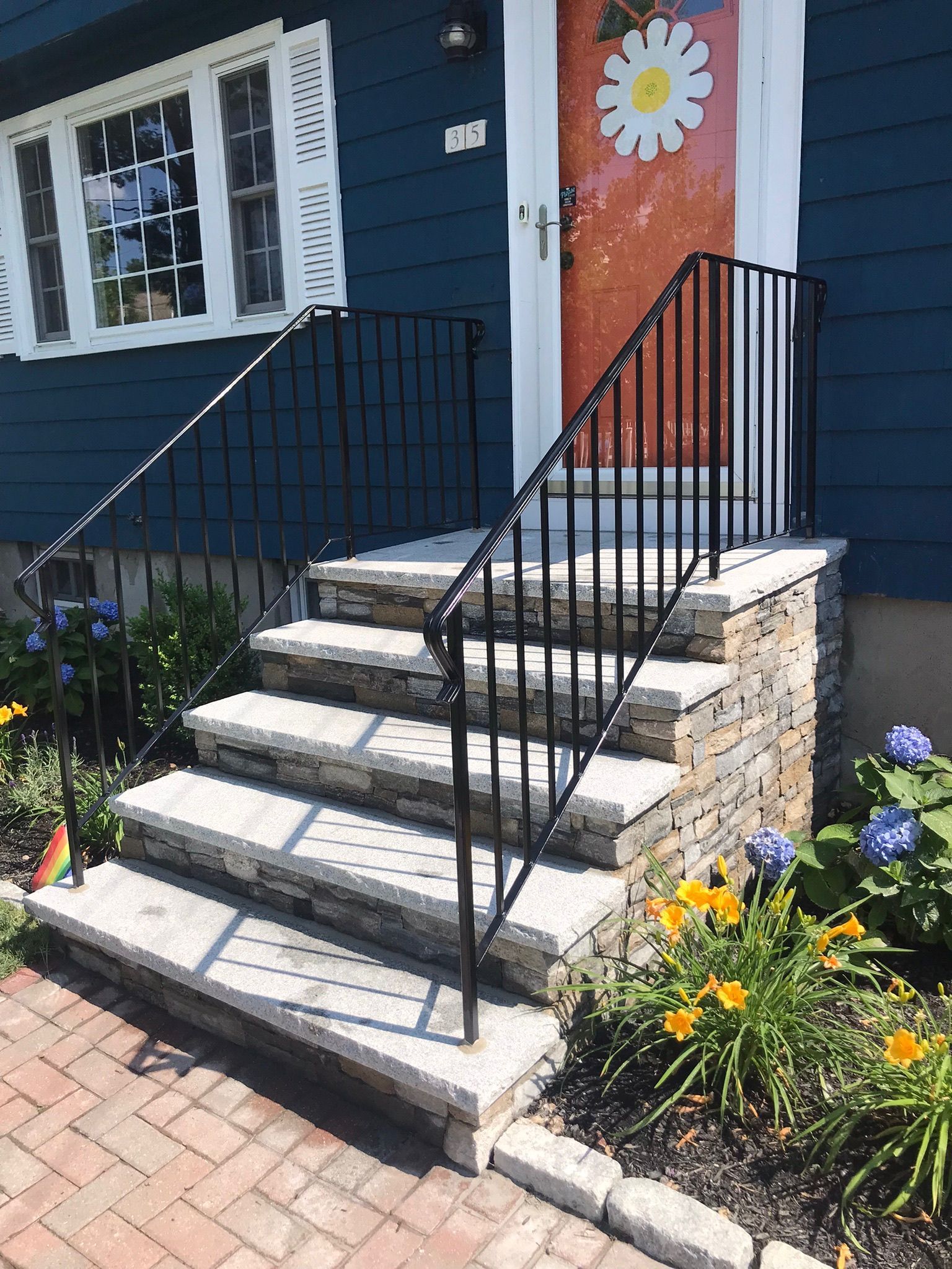 Front steps with black railings leading to a blue house door decorated with a flower mural, bordered by flowers.