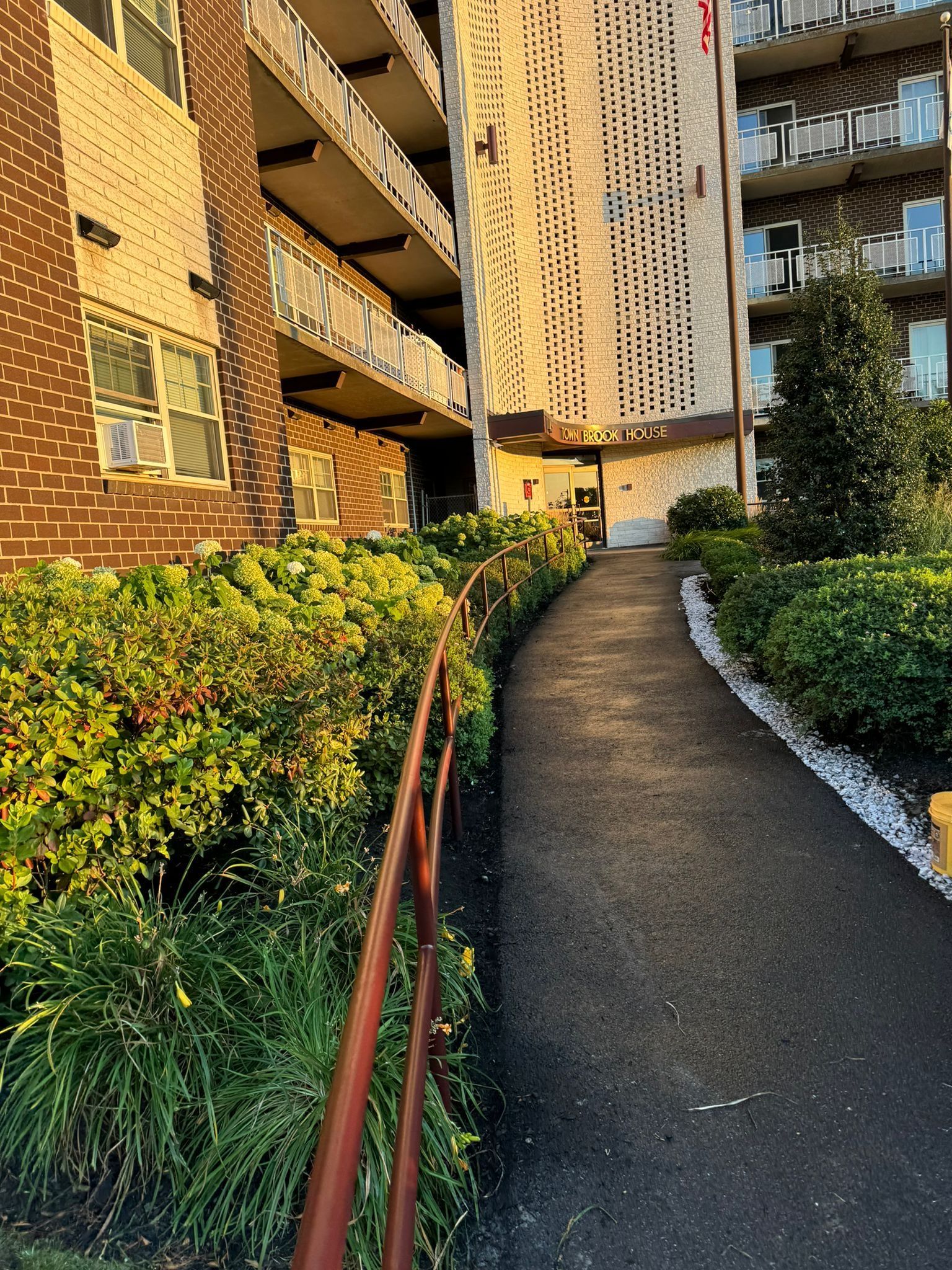 Pathway between apartment buildings with railings, greenery, and a curved accessible ramp in warm sunlight