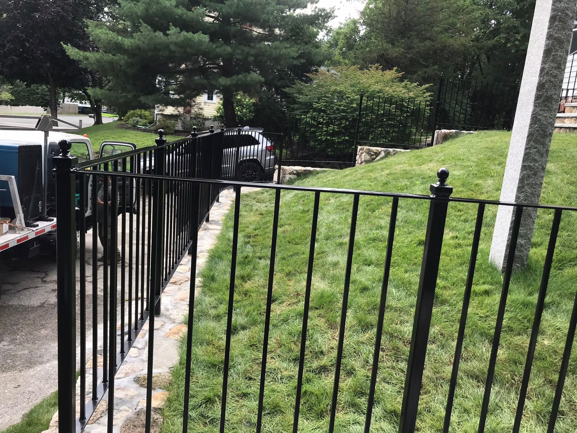 Black metal fence beside a grassy yard, with trees and a vehicle in the background.