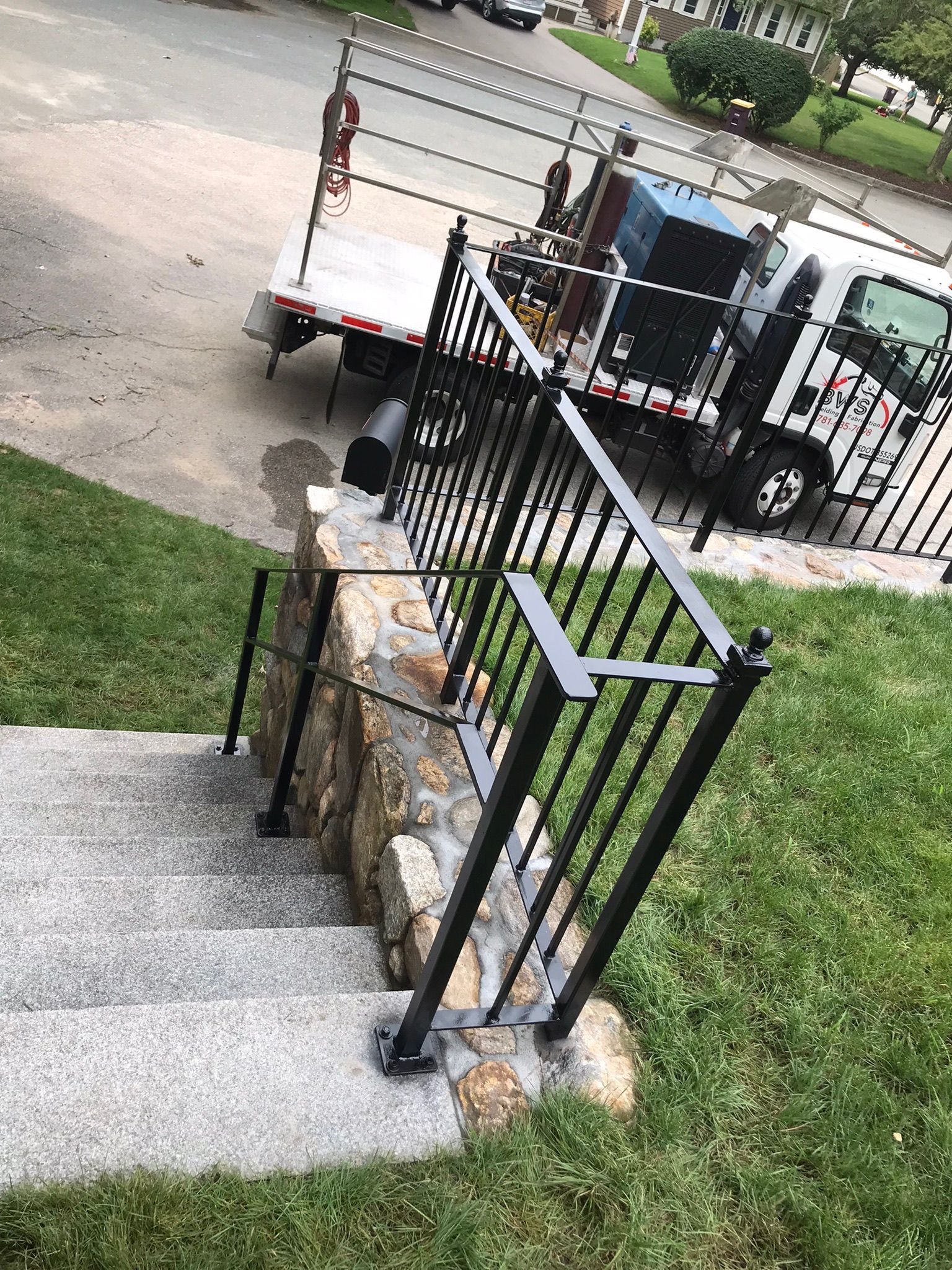 Black iron gate beside stone steps and grass, with a parked utility trailer on the street beyond.