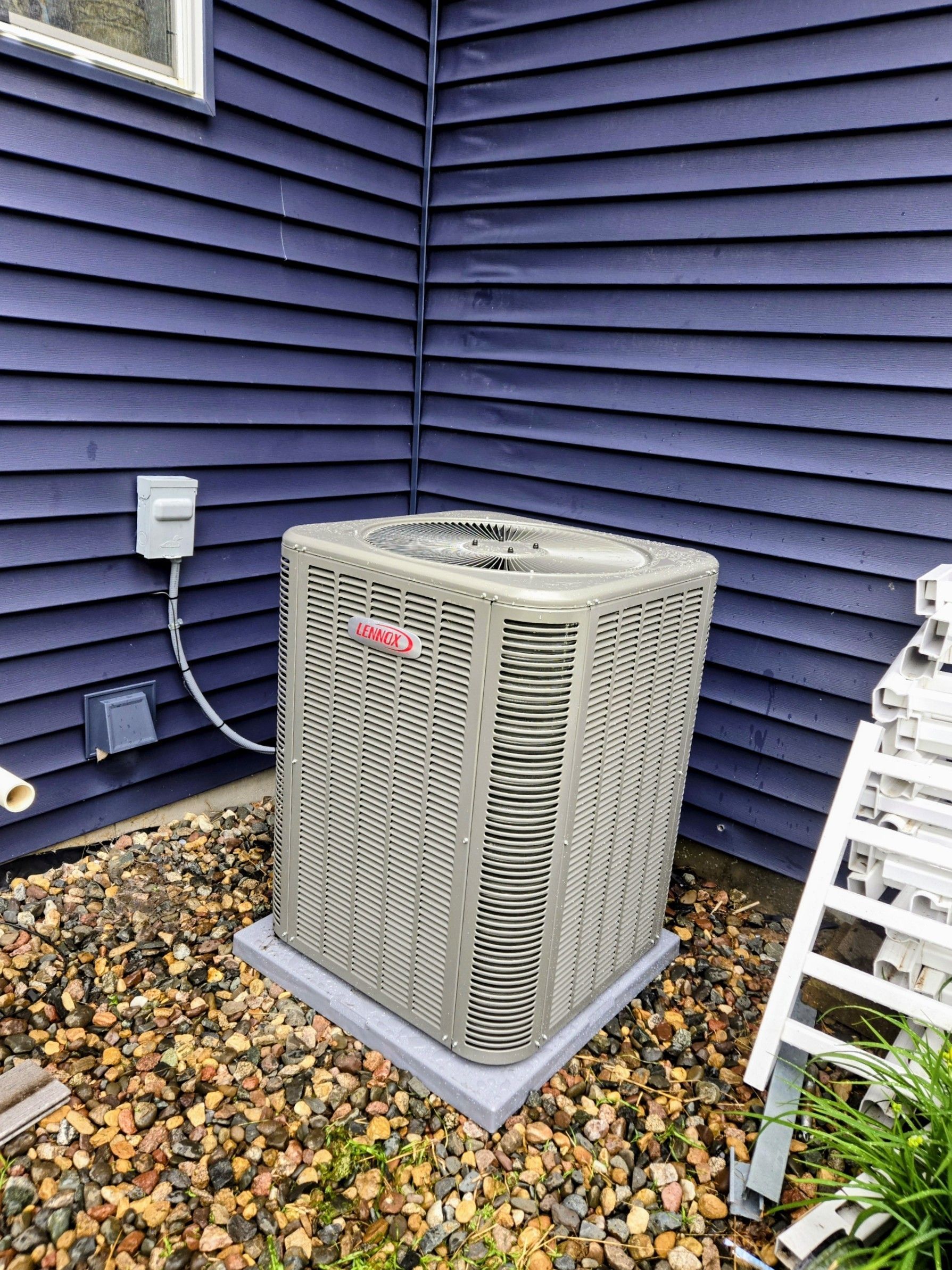 Outdoor air conditioner unit beside a blue house wall and gravel, near a white ladder