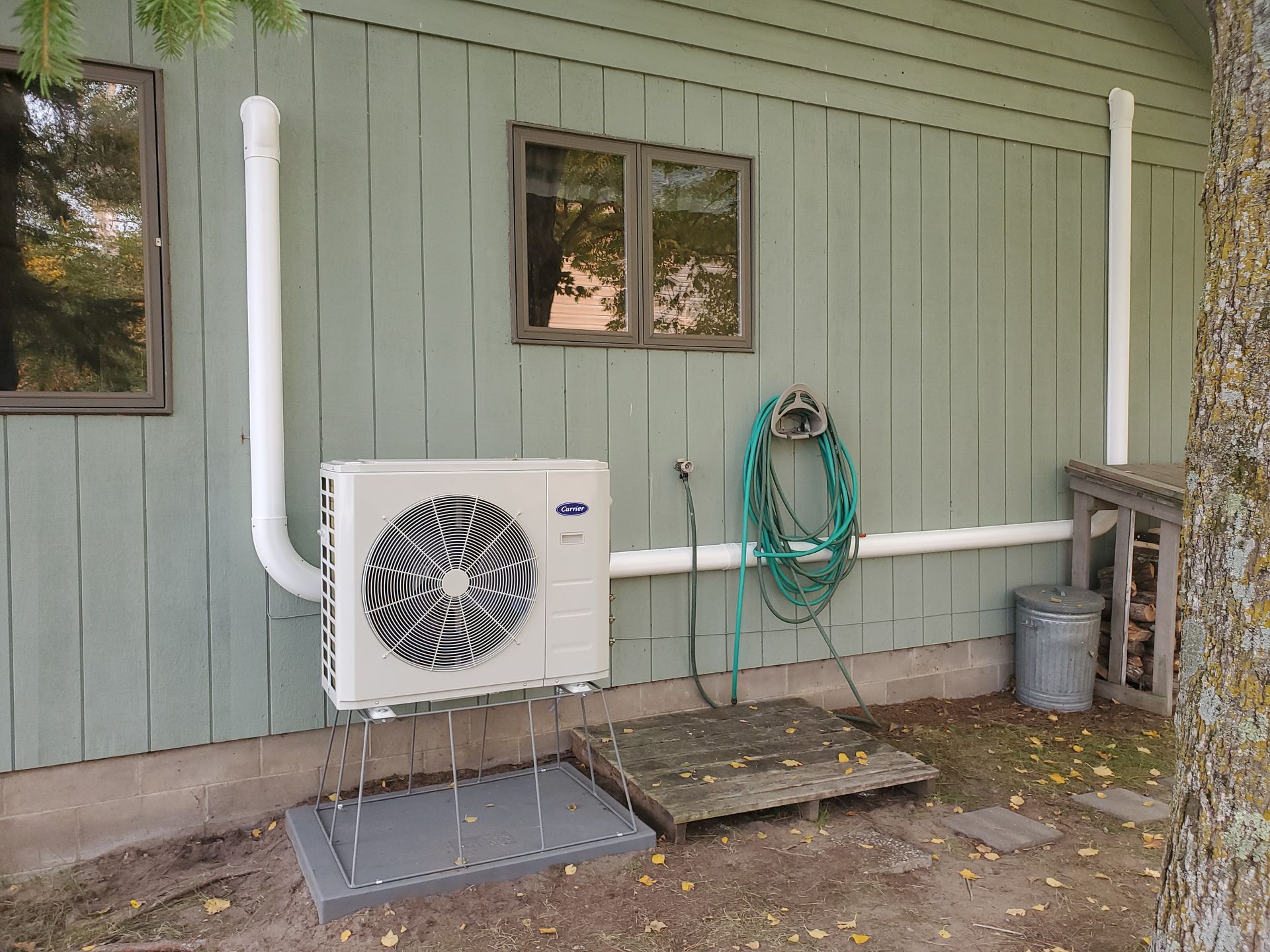 A wall-mounted heat pump unit with white piping on the exterior of a green house, next to a hose and wooden platform.