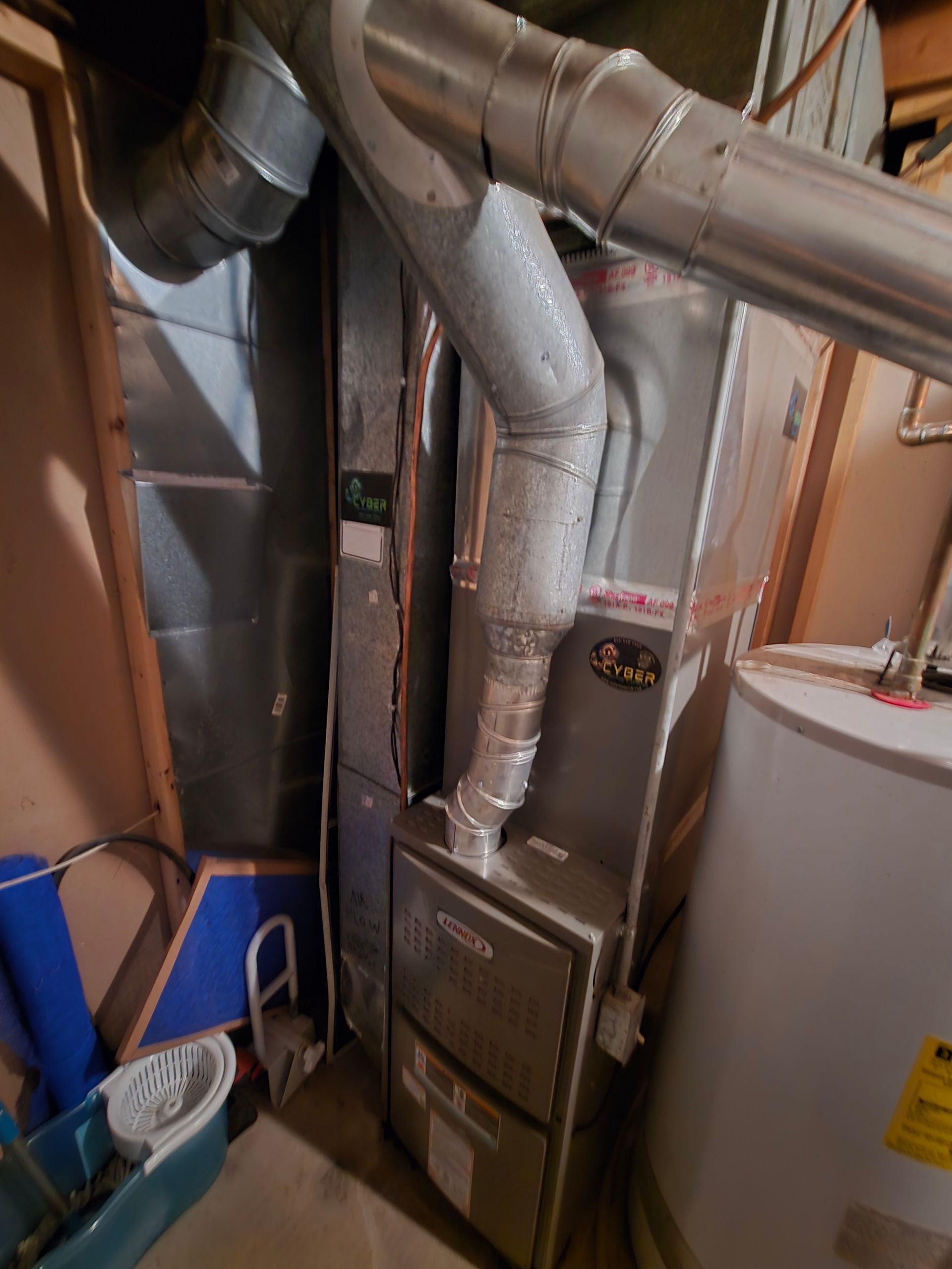 A furnace unit stands in a basement next to a water heater, with silver metal ductwork branching out from the top.