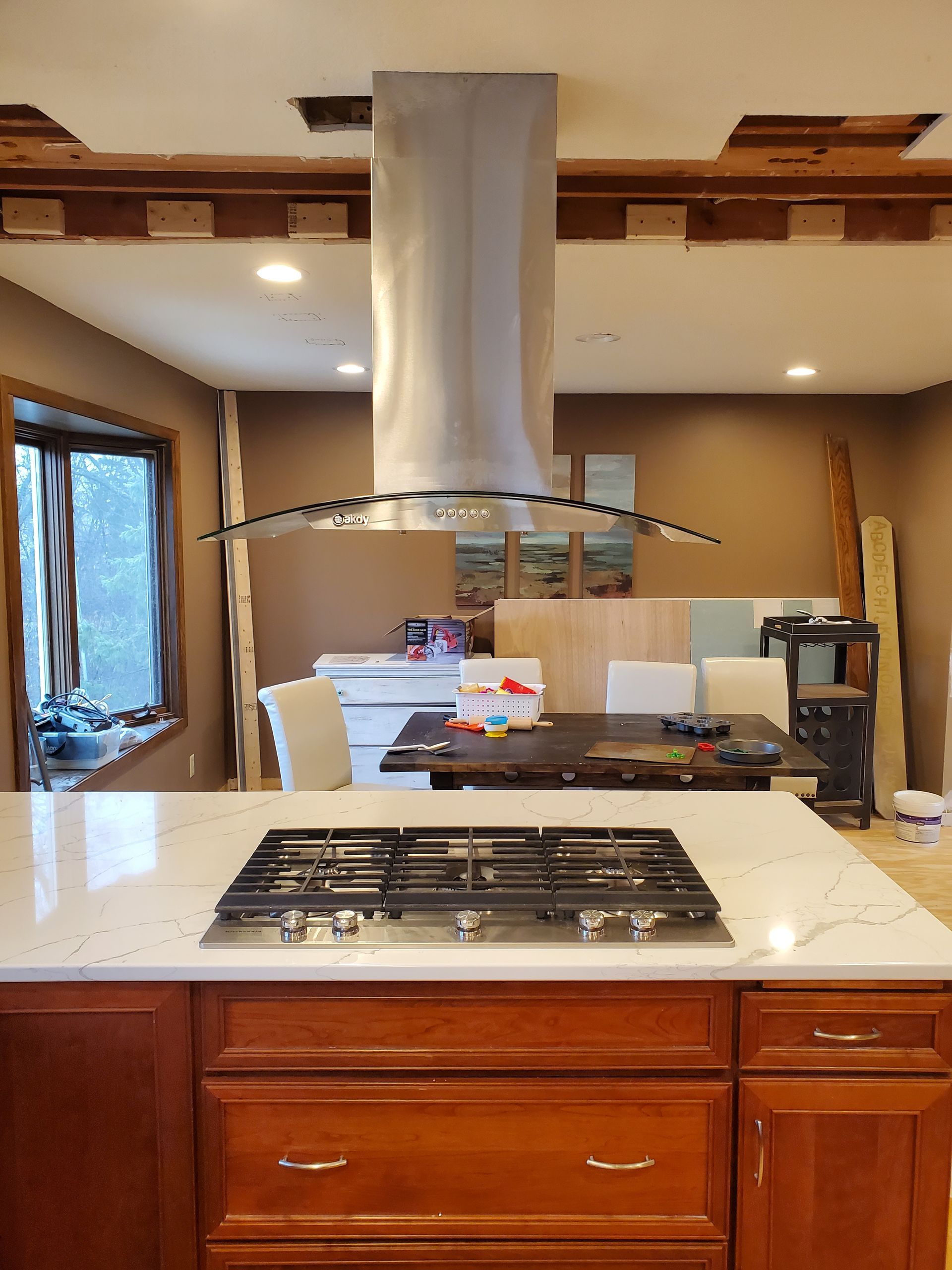 A kitchen island with a gas cooktop sits beneath a stainless steel island range hood in an unfinished ceiling space.