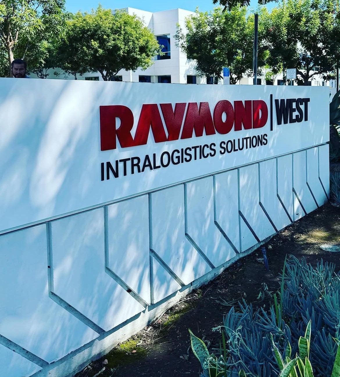 Raymond West intralogistics solutions sign on a white wall outdoors in front of trees