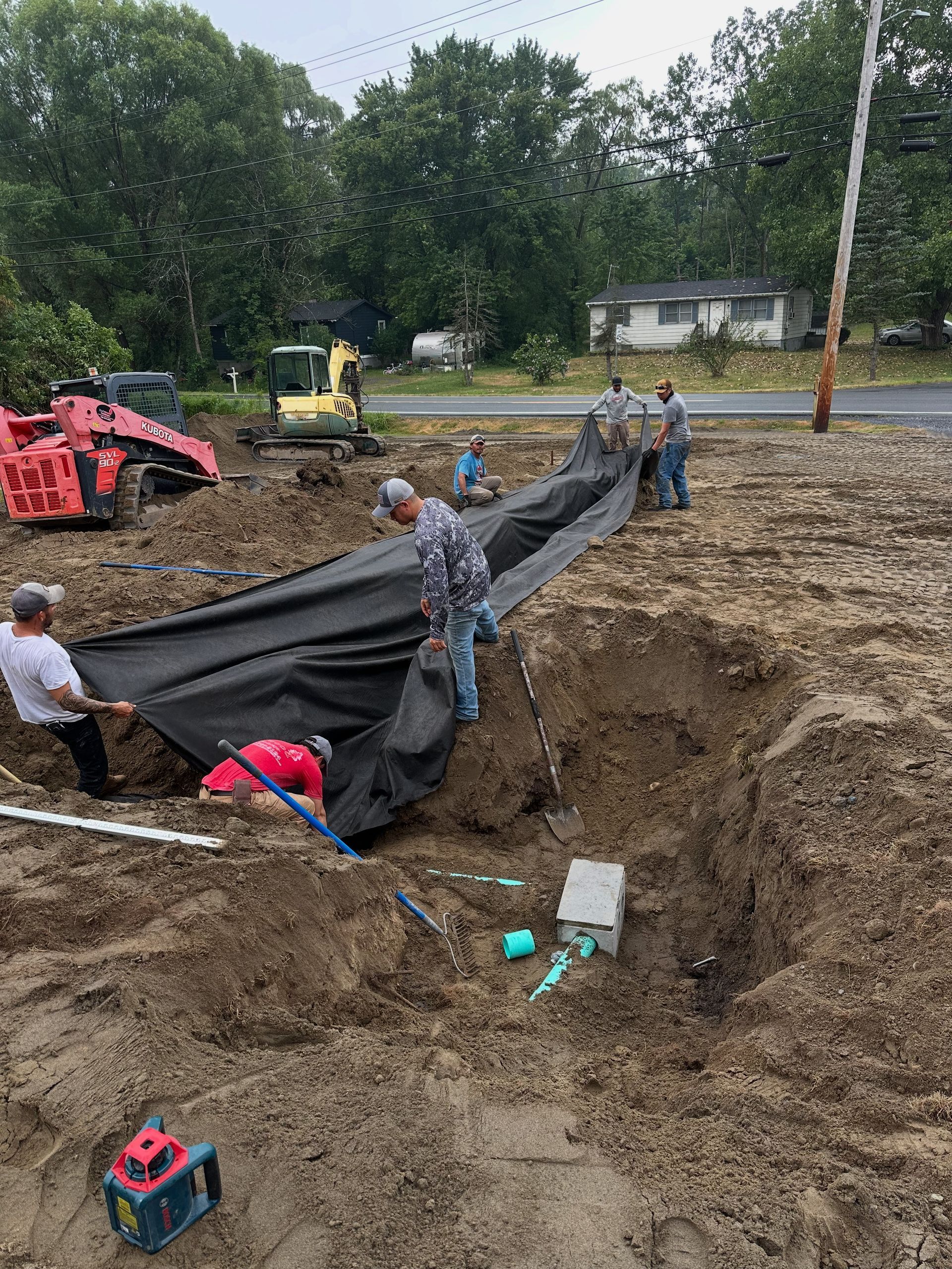 Construction workers installing a black liner in a large trench on a dirt lot with heavy machinery and blue piping visible.