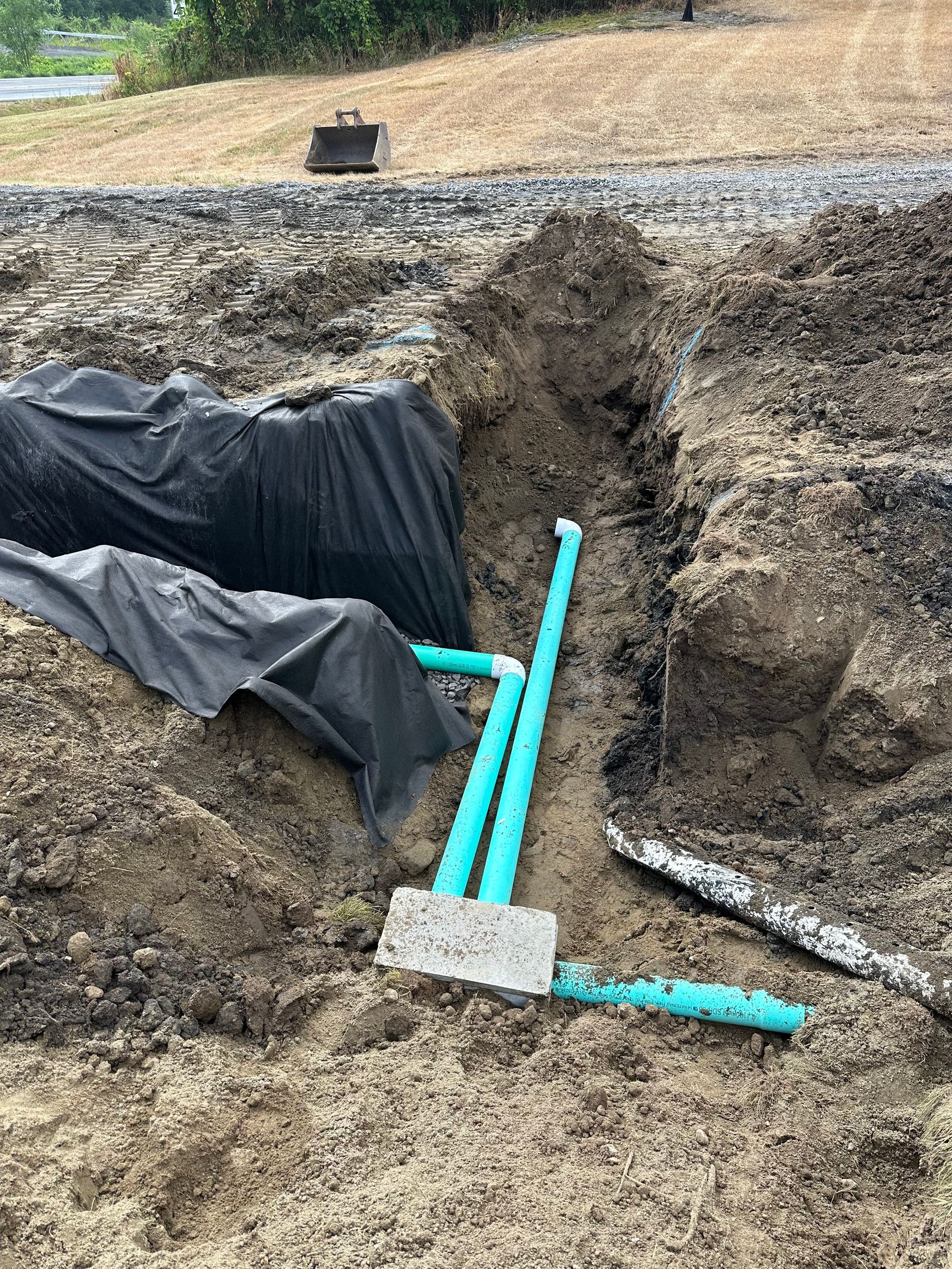 Pipes in a trench, with a concrete block and connections. A black sheet covers a dug-out area nearby.