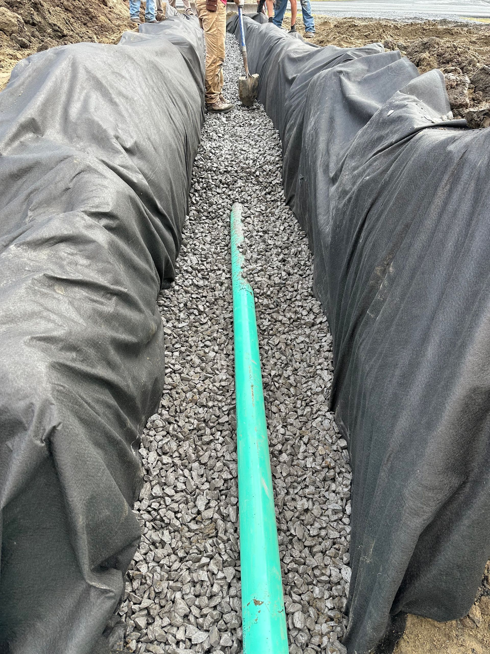 A trench lined with black fabric and filled with gravel surrounds a green pipe, likely for drainage, outdoors.