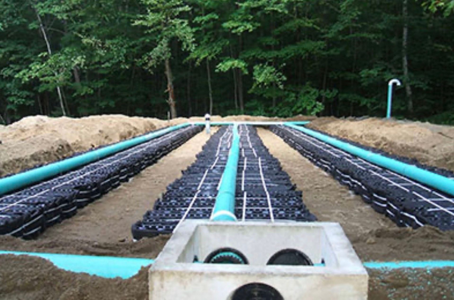 A septic system drain field with buried perforated pipes, sand, and gravel, set among trees. Blue and green pipes are prominent.