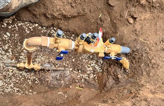 Gold-colored water valve assembly in a dirt trench, connected to pipes and gravel.