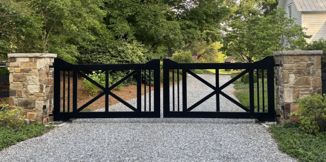 Stainless steel driveway gate, vertical bars, opening onto driveway with greenery and blue sky.