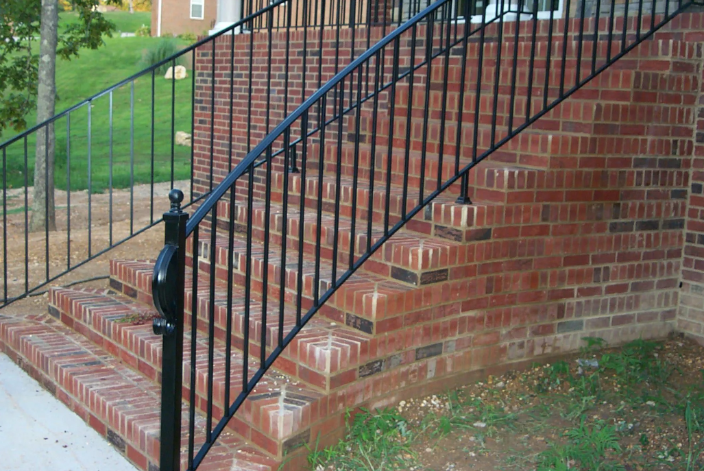 Elegant staircase with ornate black railing, leading to a dining area with round table, green chairs, and floral arrangement.