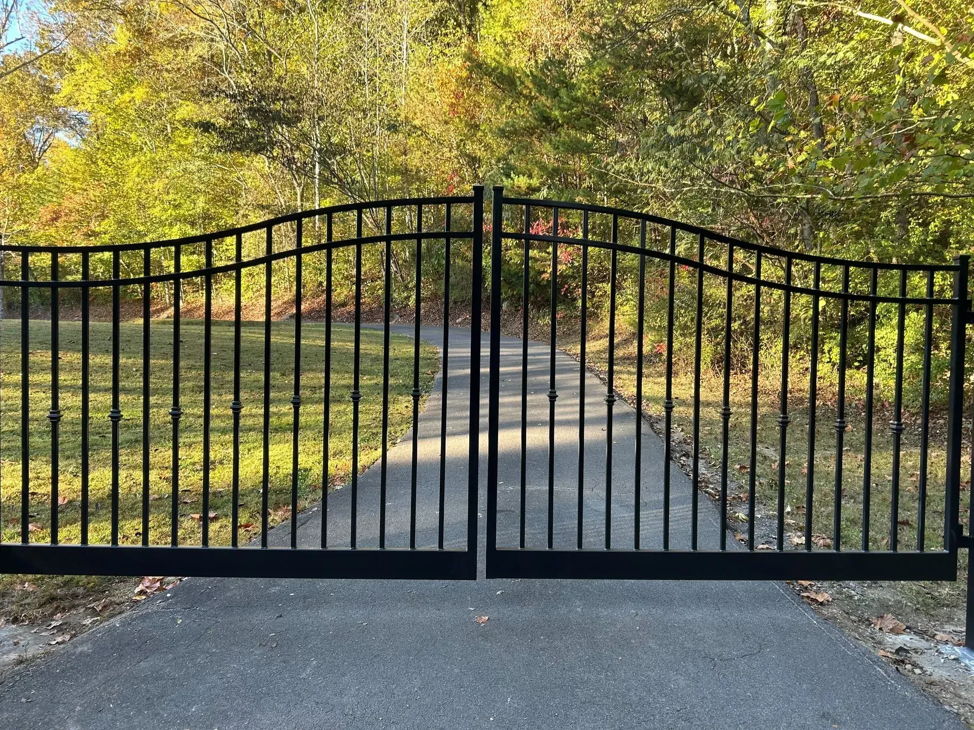 Black metal driveway gates, closed, on a paved path leading into a wooded area.