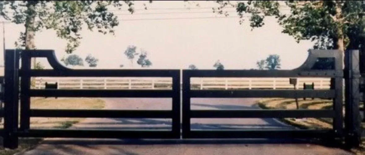 Wooden gate on a paved road, framed by trees. A white fence is visible in the background.