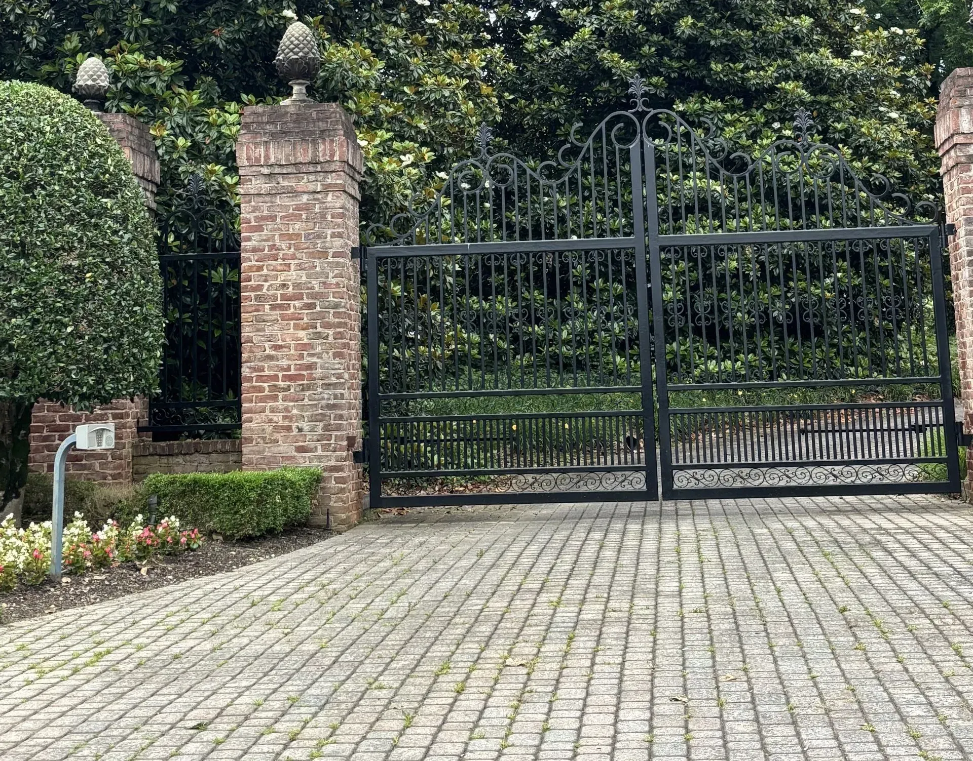Black wrought iron gates between brick pillars on a cobblestone driveway.