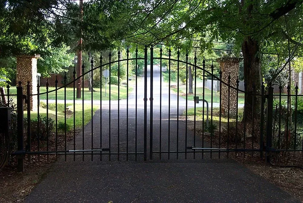 Black iron gates open to a driveway lined with trees.