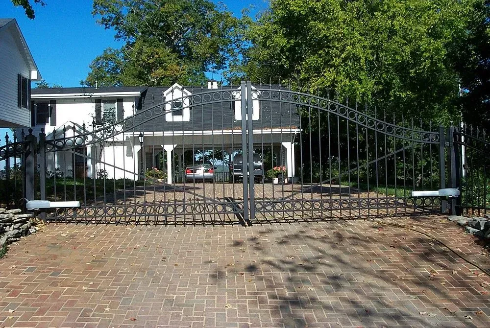 Black wrought iron gate in front of a white house with a driveway and cars under a carport.
