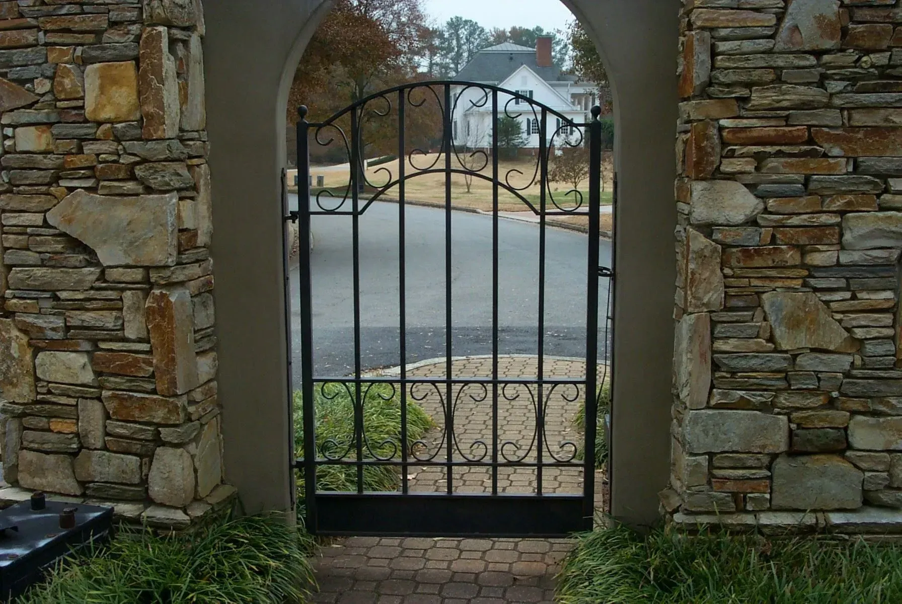 Black wrought iron gate set within stone archway, leading to a street with houses.