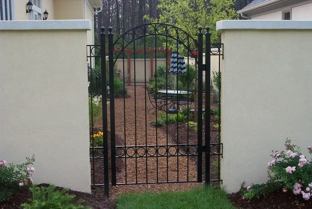 Black iron gate in a beige stucco wall, leading to a garden path with plants and trees.