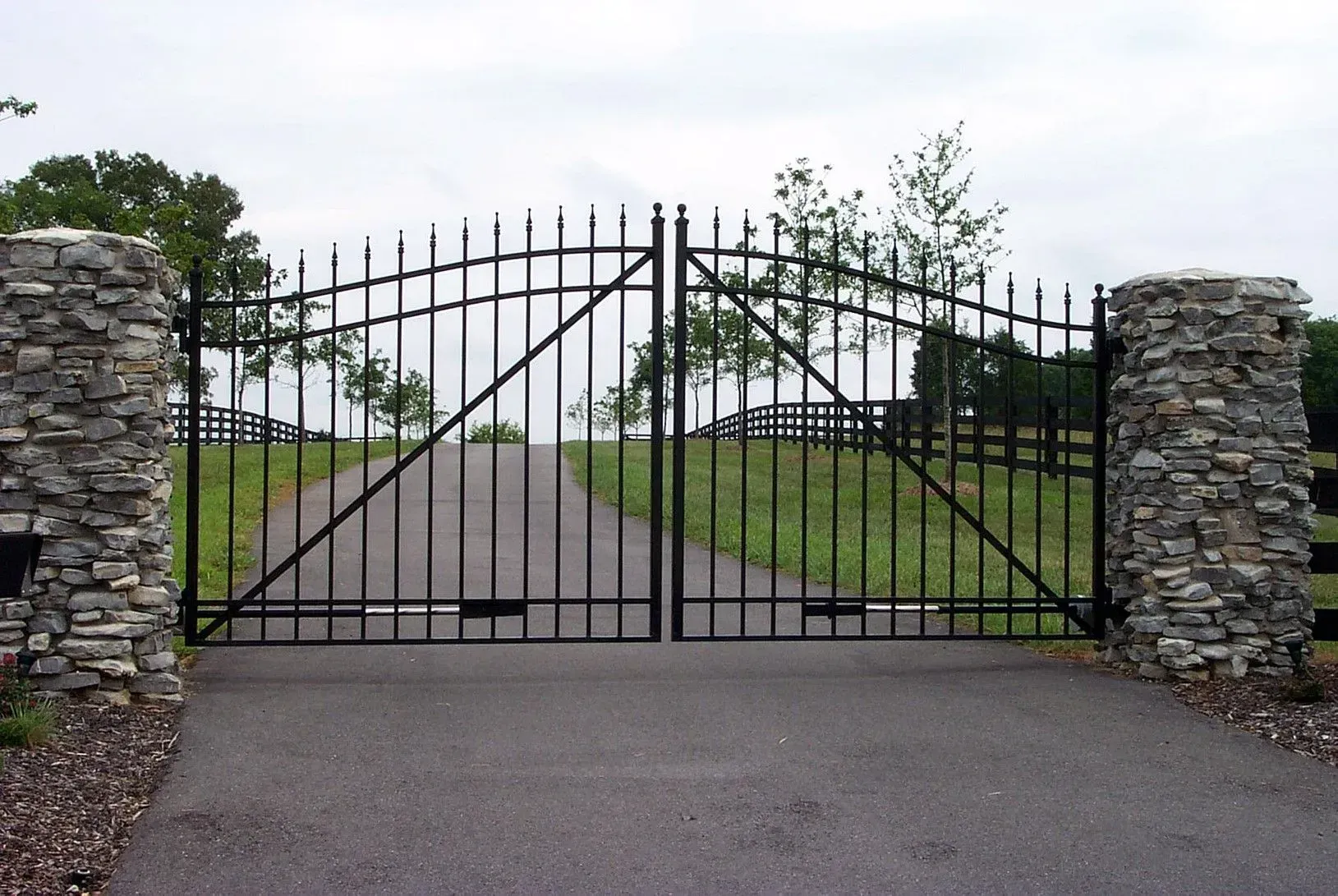 Black metal gates between stone columns open to a paved driveway, leading up a slight hill.