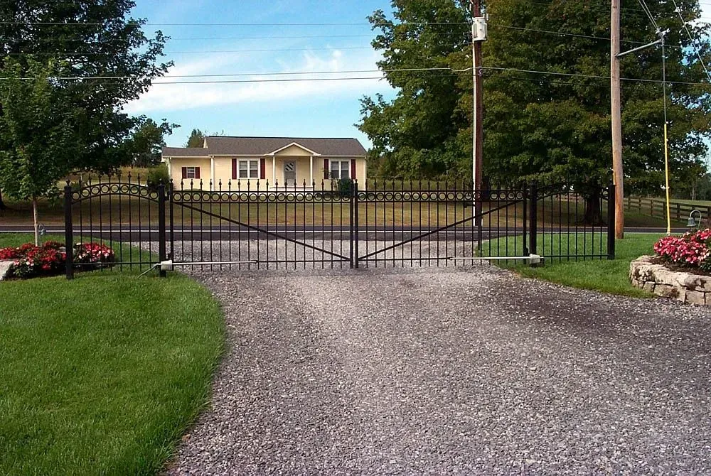 Black iron gates open to a gravel driveway leading to a yellow house with a dark roof; green lawn and trees surround it.