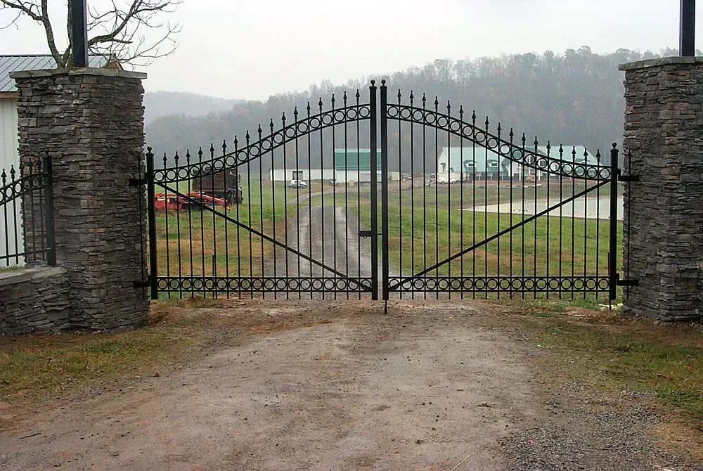 Black metal gates between stone pillars, leading to a dirt road. Green fields and buildings in the background.